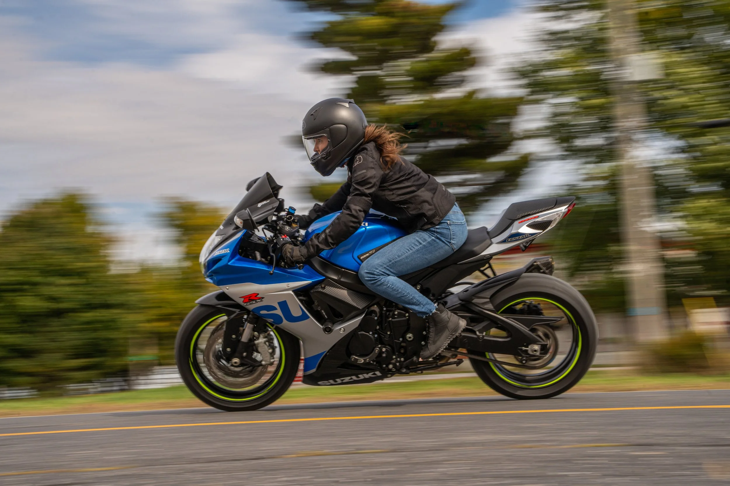Une personne portant un casque noir, une veste noire et des jeans bleus, qui roule rapidement sur une moto sport bleu avec des accents noirs et des pneus à jantes vertes, en déplacement sur une route en milieu urbain ou rural avec des arbres flous en