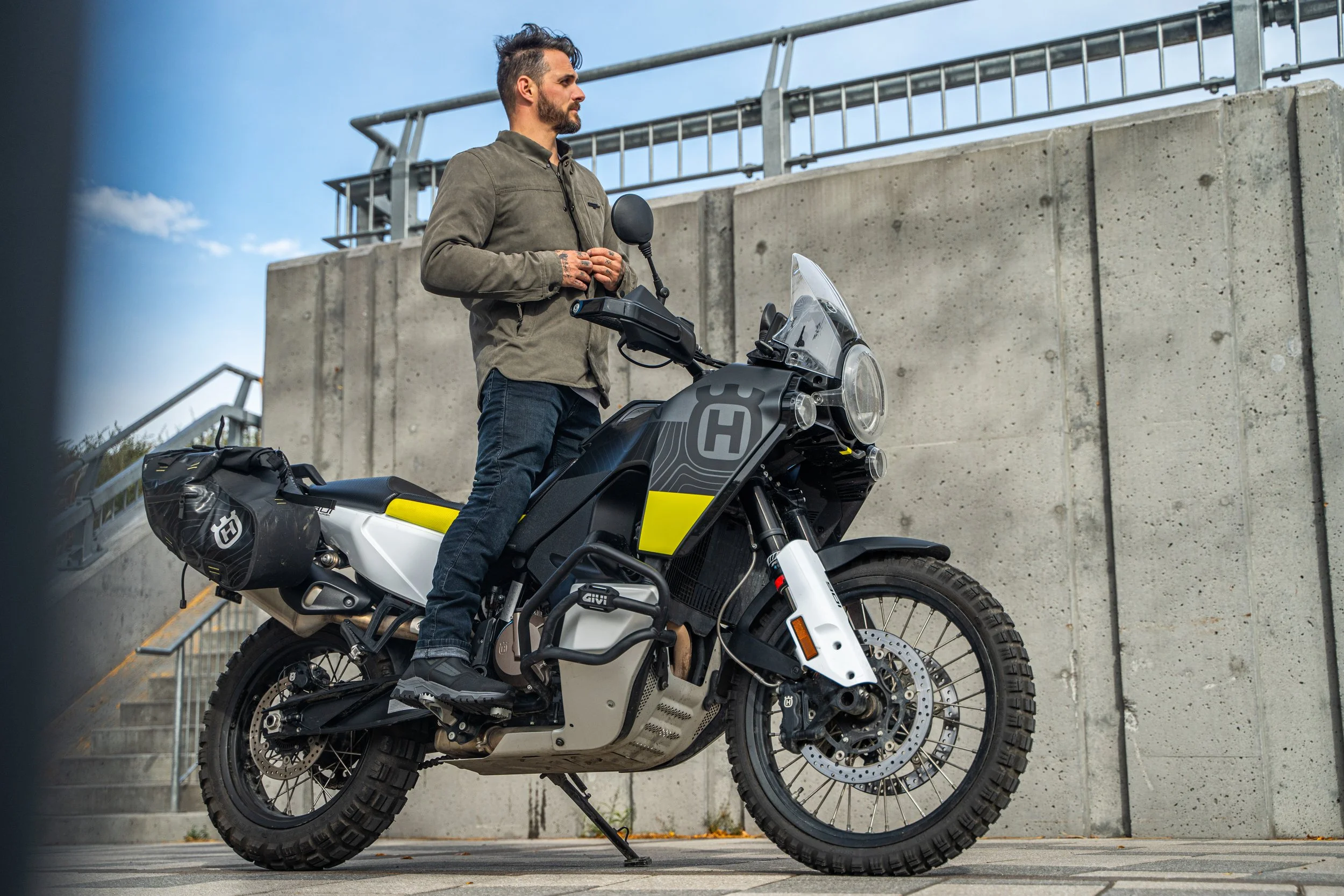 Un homme avec une barbe se tient debout sur une moto noire et jaune, en regardant vers la gauche, en dehors d'un mur en béton, par temps ensoleillé.