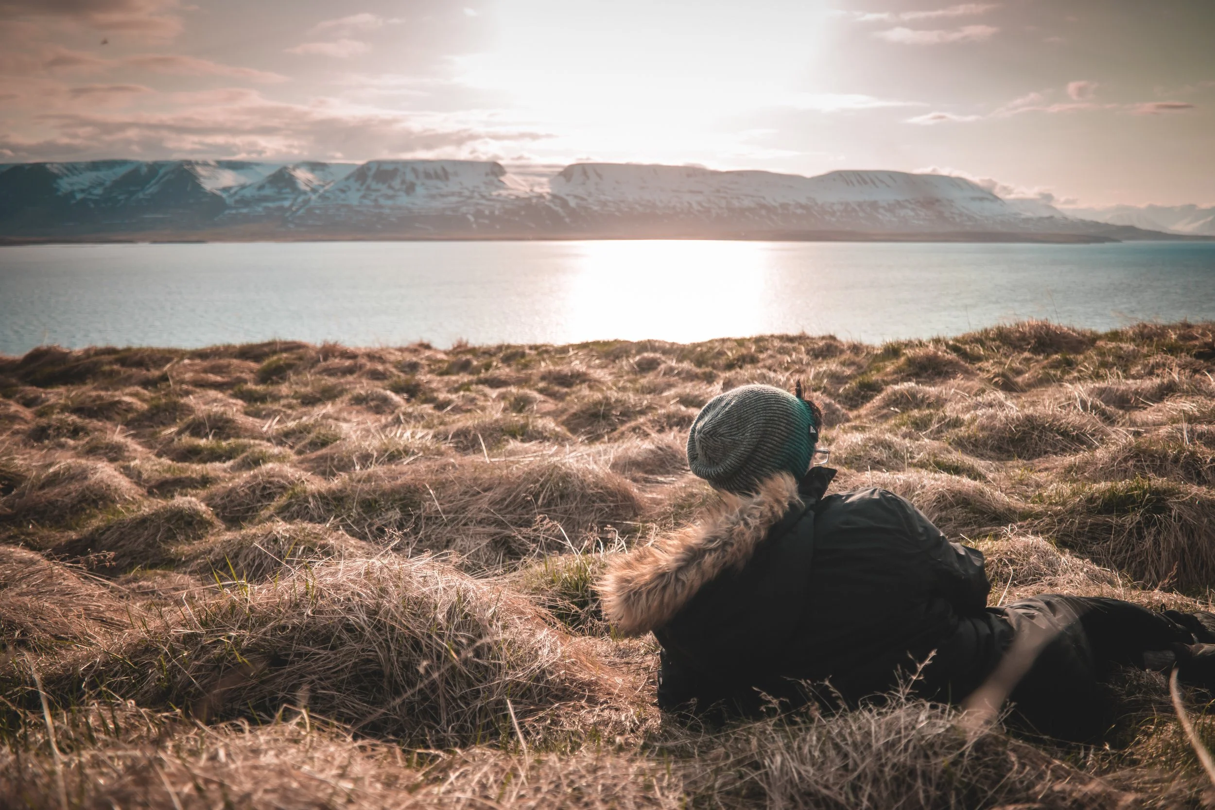Une personne allongée dans une prairie regarde un lac avec des montagnes enneigées au loin, le soleil bas dans le ciel.