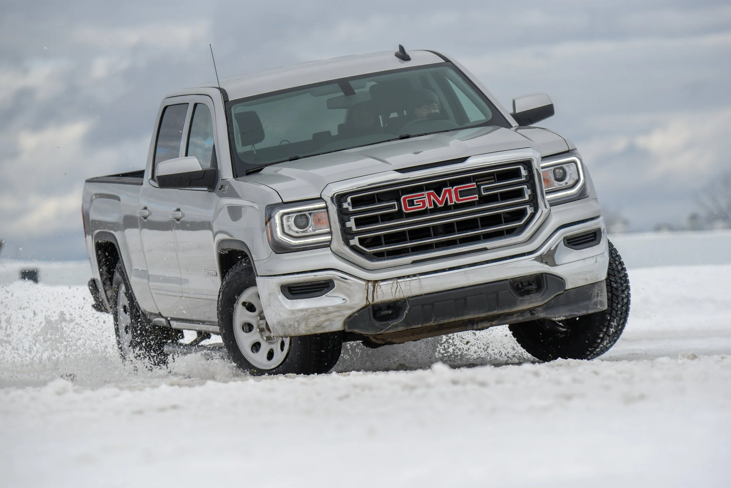 Une camionnette blanche GMC en mouvement sur un terrain enneigé, avec des nuages gris en arrière-plan.