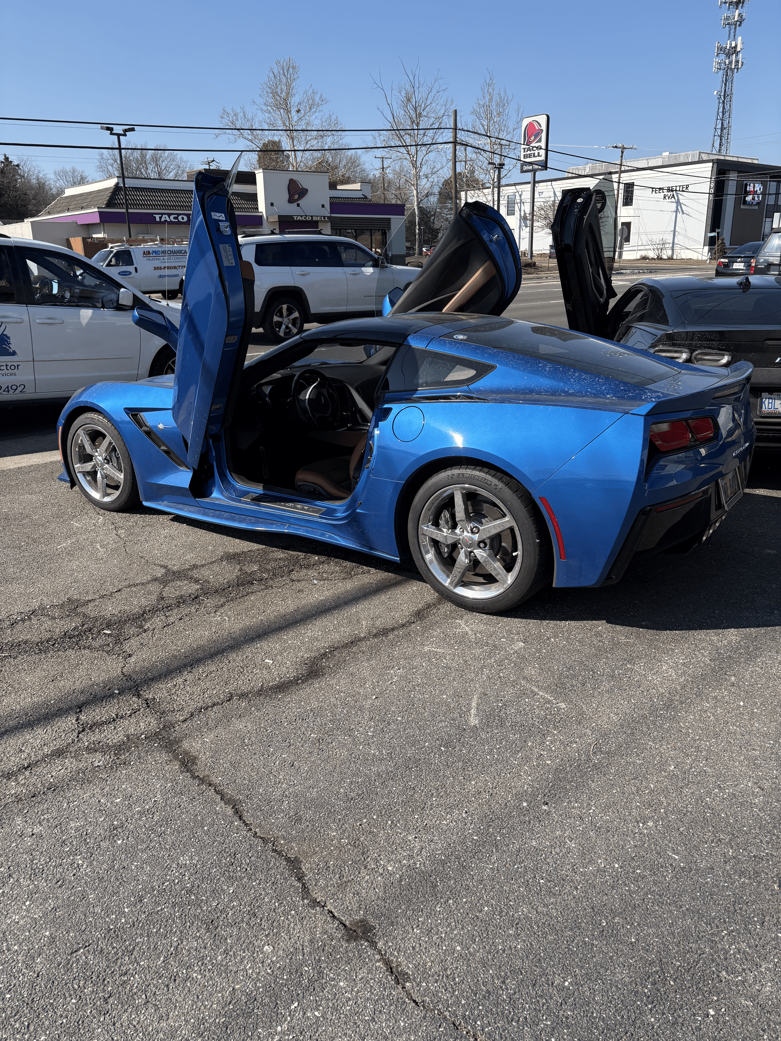 A blue sports car with gull-wing doors open parked in a lot, with other cars and a Taco Bell restaurant in the background.