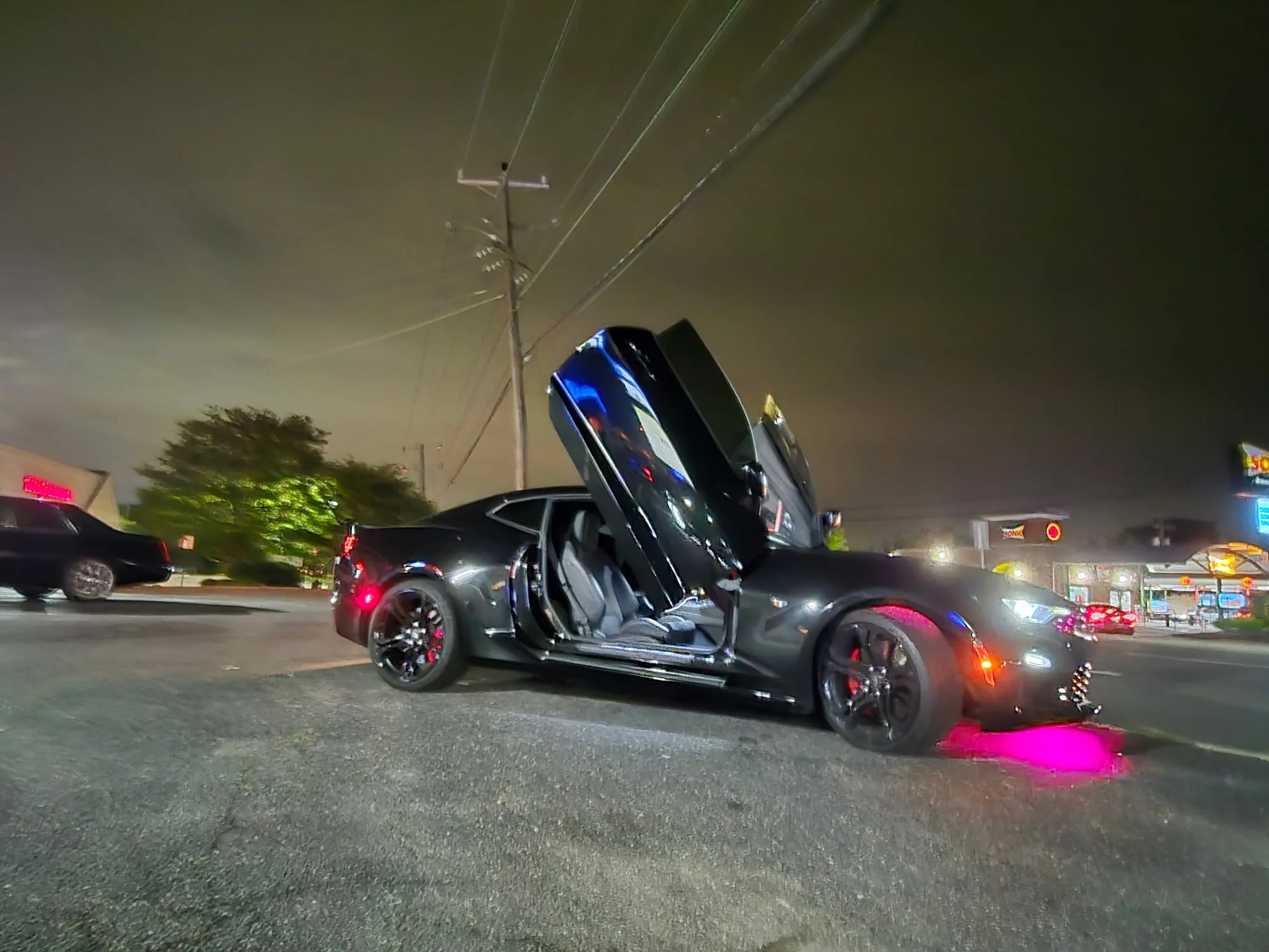 A black sports car with gullwing doors open parked on a street at night, with pink underglow lighting beneath it and parked cars, trees, utility poles, and a shopping plaza in the background.