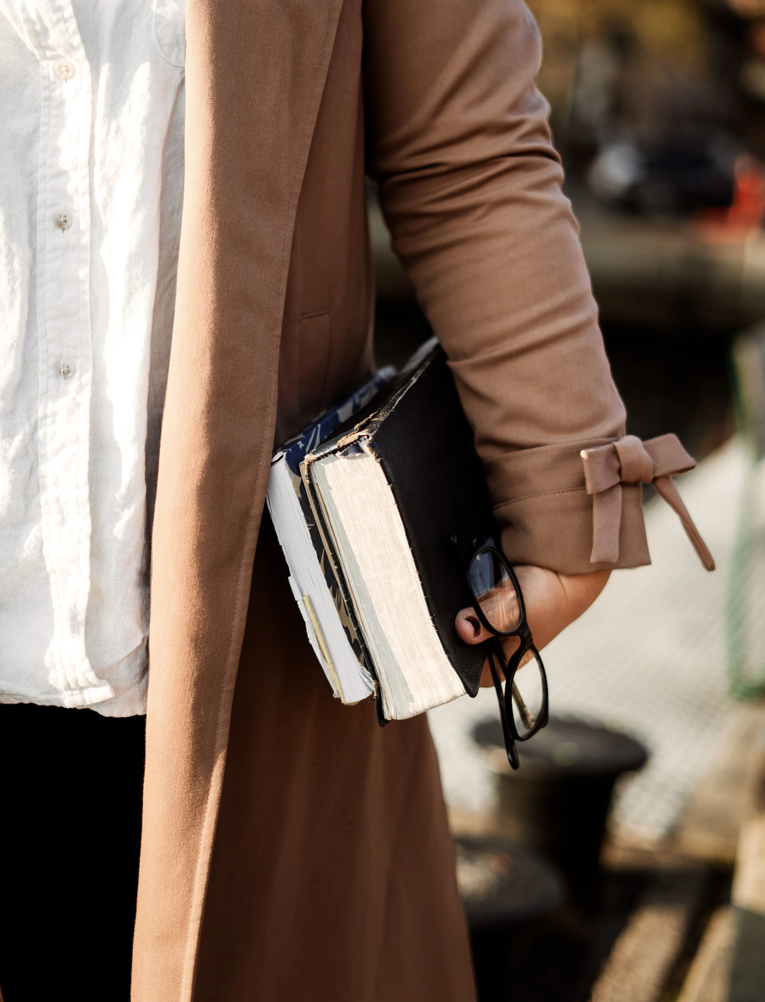 KJ from Ezer Copy Studio holding a Bible, journal and Copywriting how-to with glasses hanging from the bottom, wearing a brown coat and white shirt.