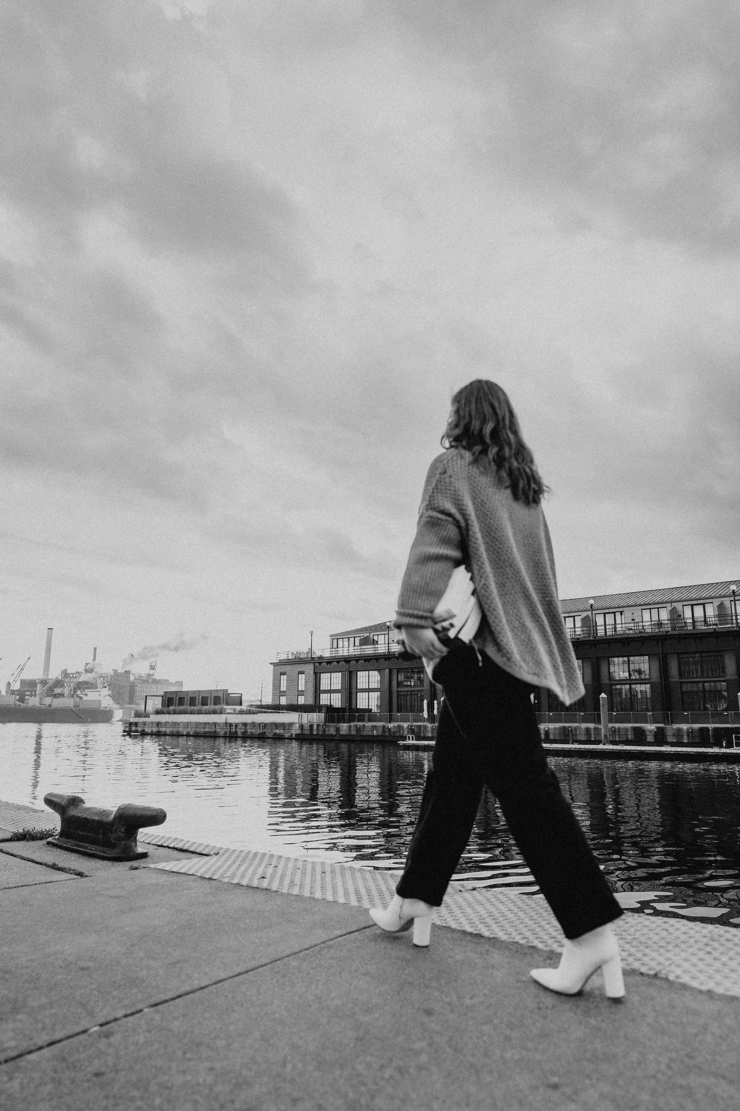 KJ from Ezer Copy Studio walking along the Fells Point waterfront in Baltimore City on a cloudy day, carrying a laptop under her arm, wearing a sweater, black pants, and white heeled boots.