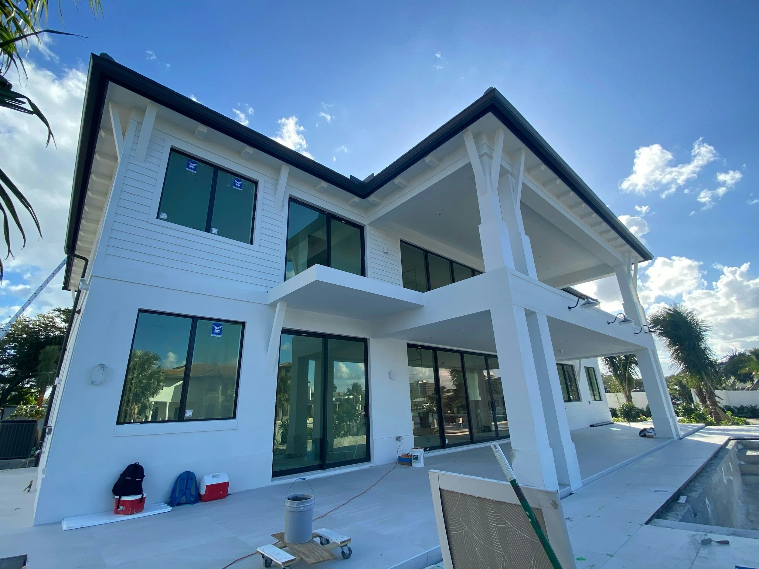 Modern white house with large glass windows, under construction, with scaffolding and construction tools on patio, surrounded by palm trees, under blue sky with clouds.