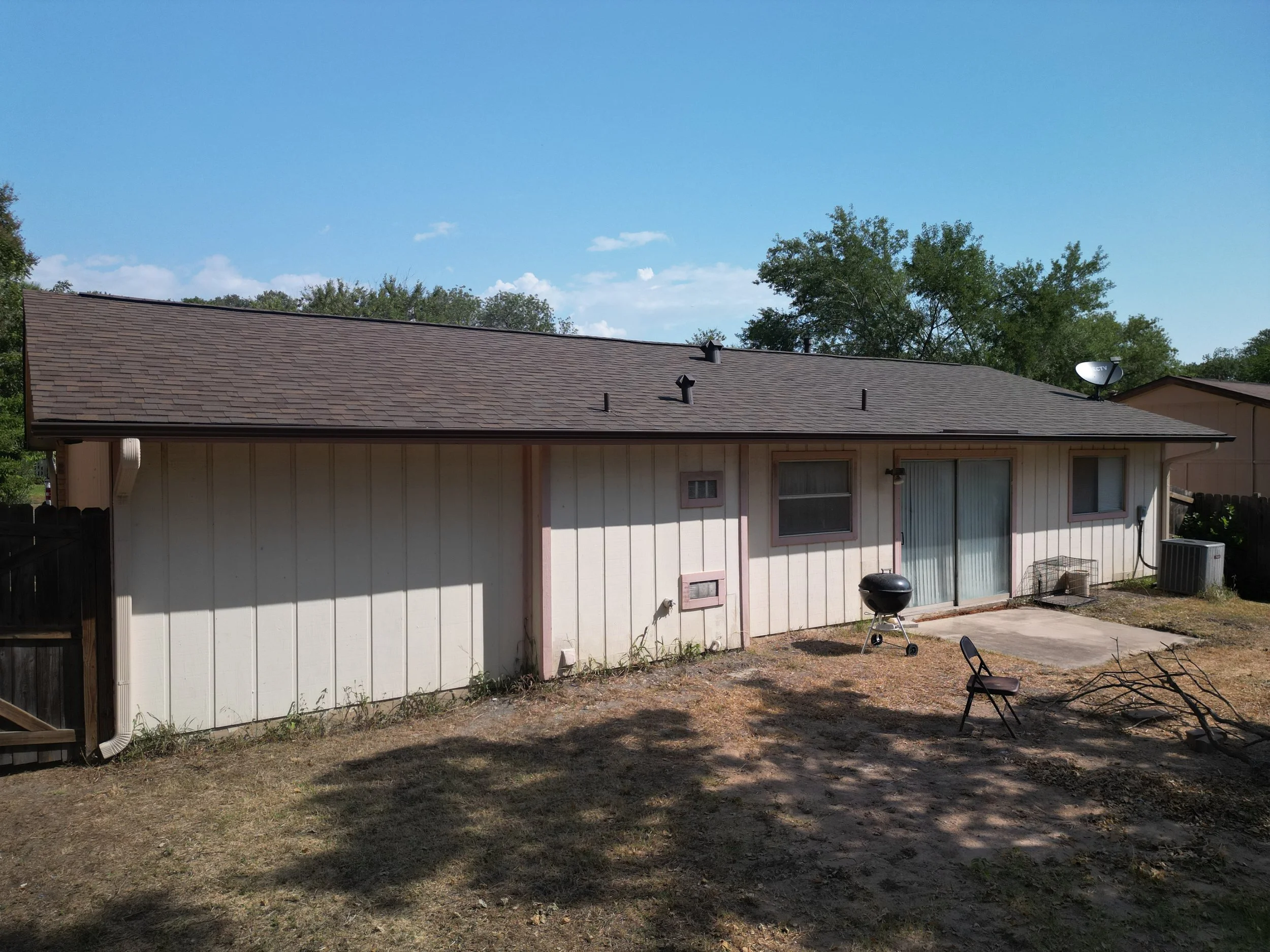 The back of a single-story house with a brown shingle roof, white wall siding, sliding glass door, and small patio area. There is a charcoal grill, a lawn chair, and a small patch of dirt in the yard under a clear blue sky.