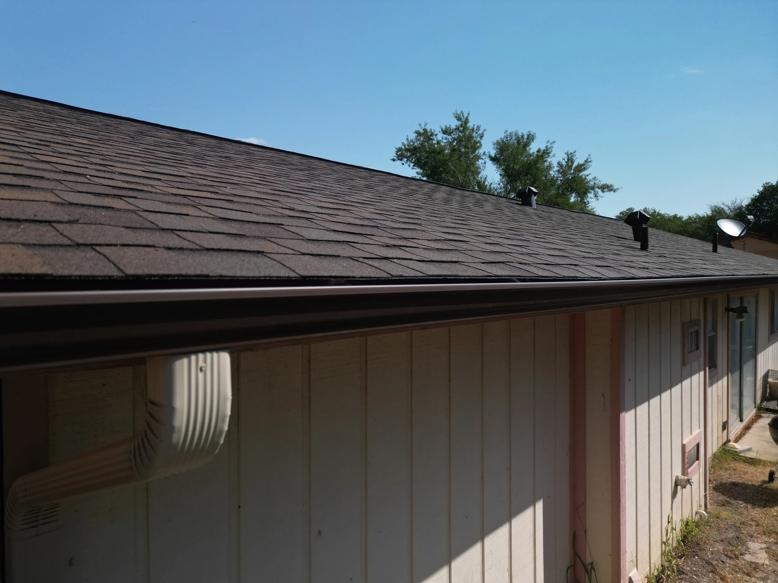 Side view of a house roof with brown shingles, a gutter, exhaust vents, and a satellite dish against a blue sky with trees in the background.