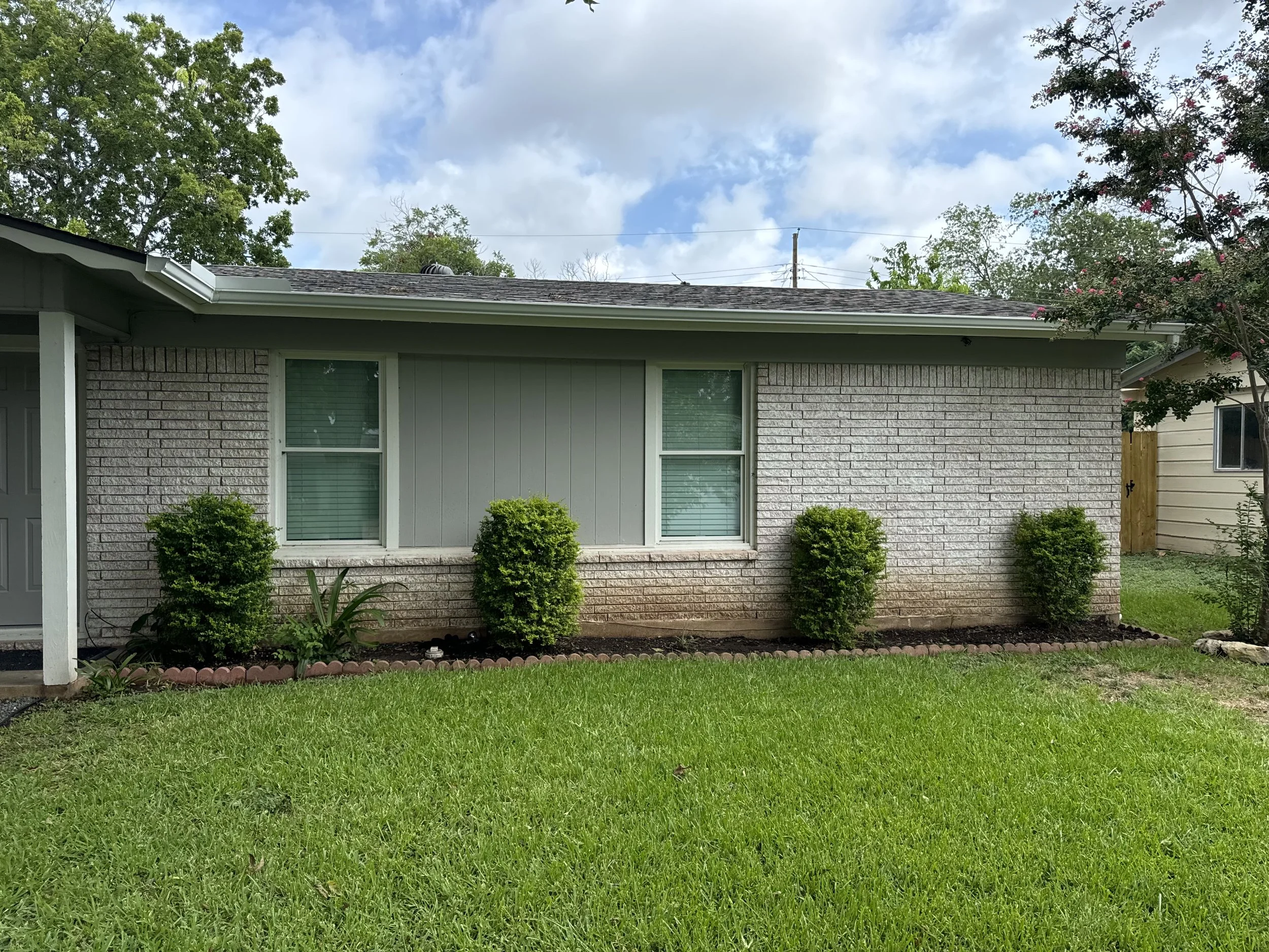 Front yard view of a house with gray brick and siding, three bushes in front, green grass, partly cloudy sky, and a tree on the right.
