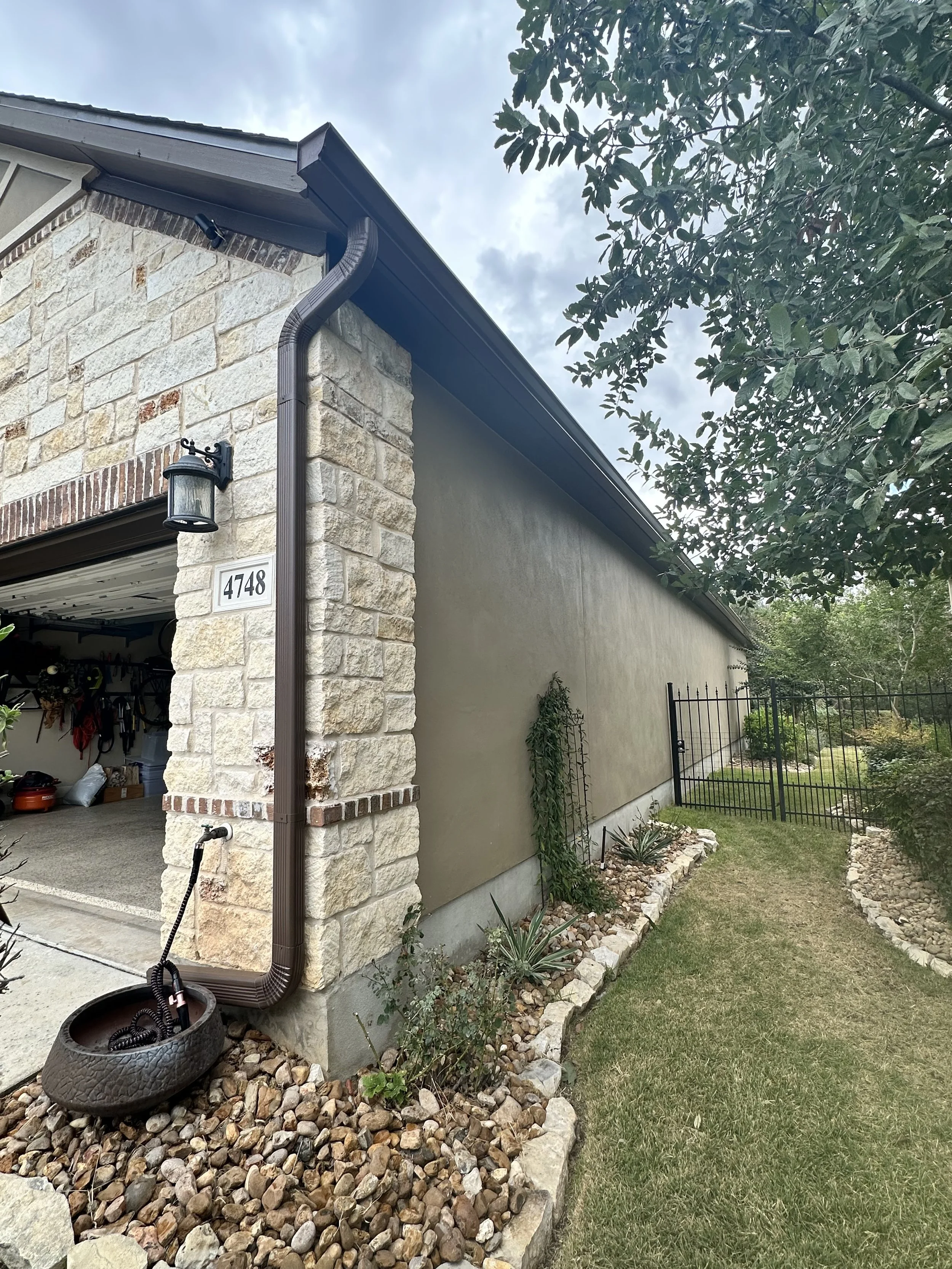 Side of a house with beige stone and stucco exterior, a brown downspout, a garage door partially visible, and a garden with rocks and plants, next to a black metal fence.