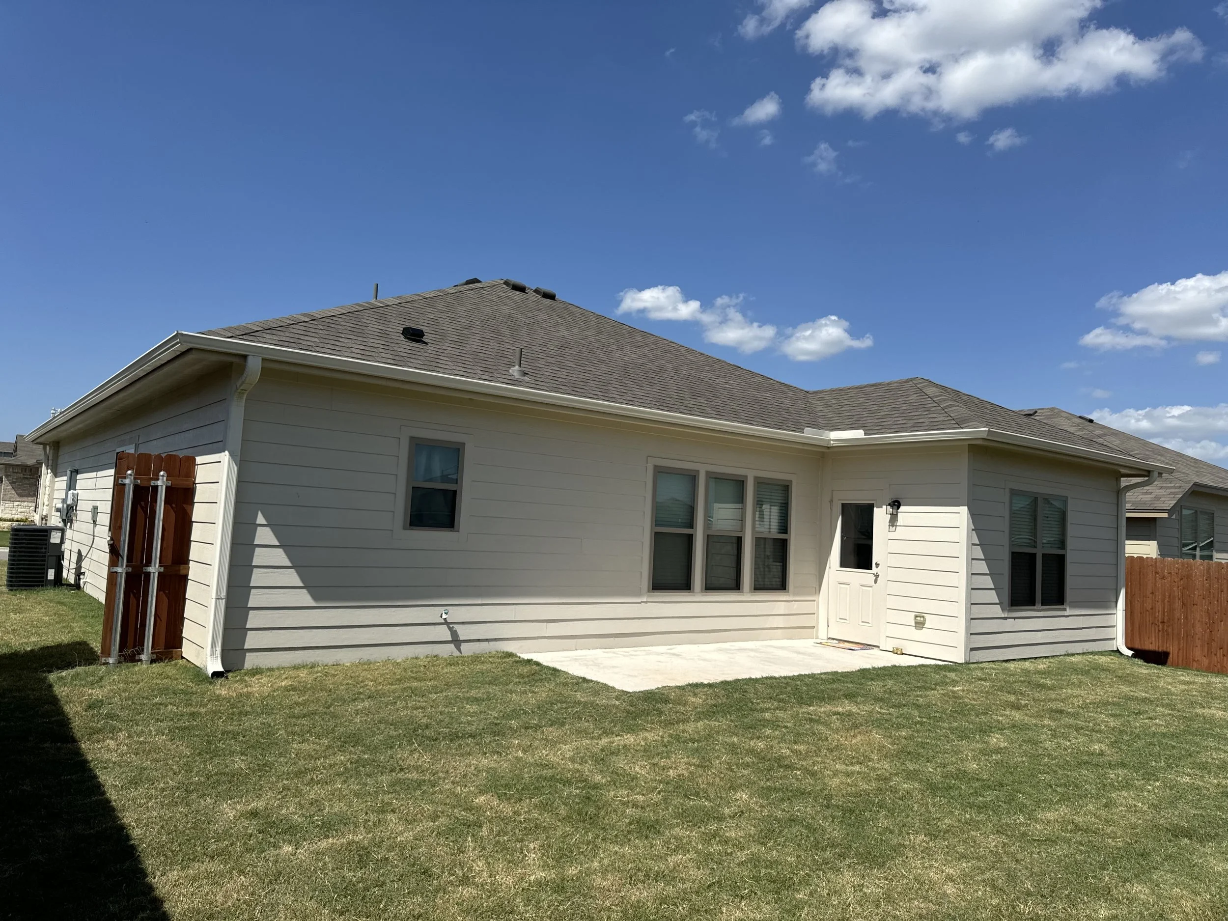 Backyard view of a single-story house with white siding, a brown shingle roof, four windows, a white door with a small window, a concrete patio, and a green lawn under blue sky with scattered clouds.