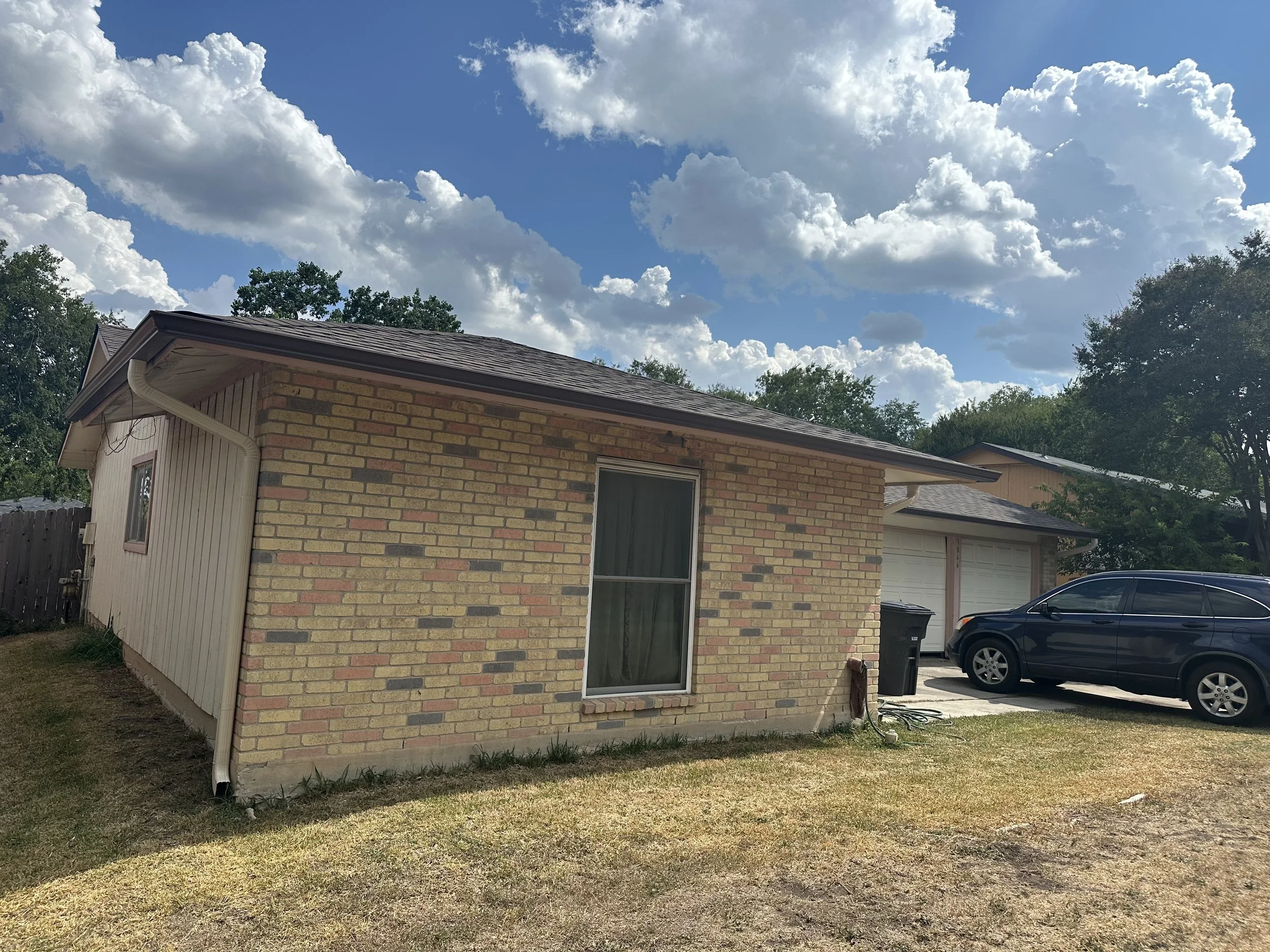 Exterior view of a brick house with a garage, parked black SUV, trash bins, and a paved driveway, under a partly cloudy sky.