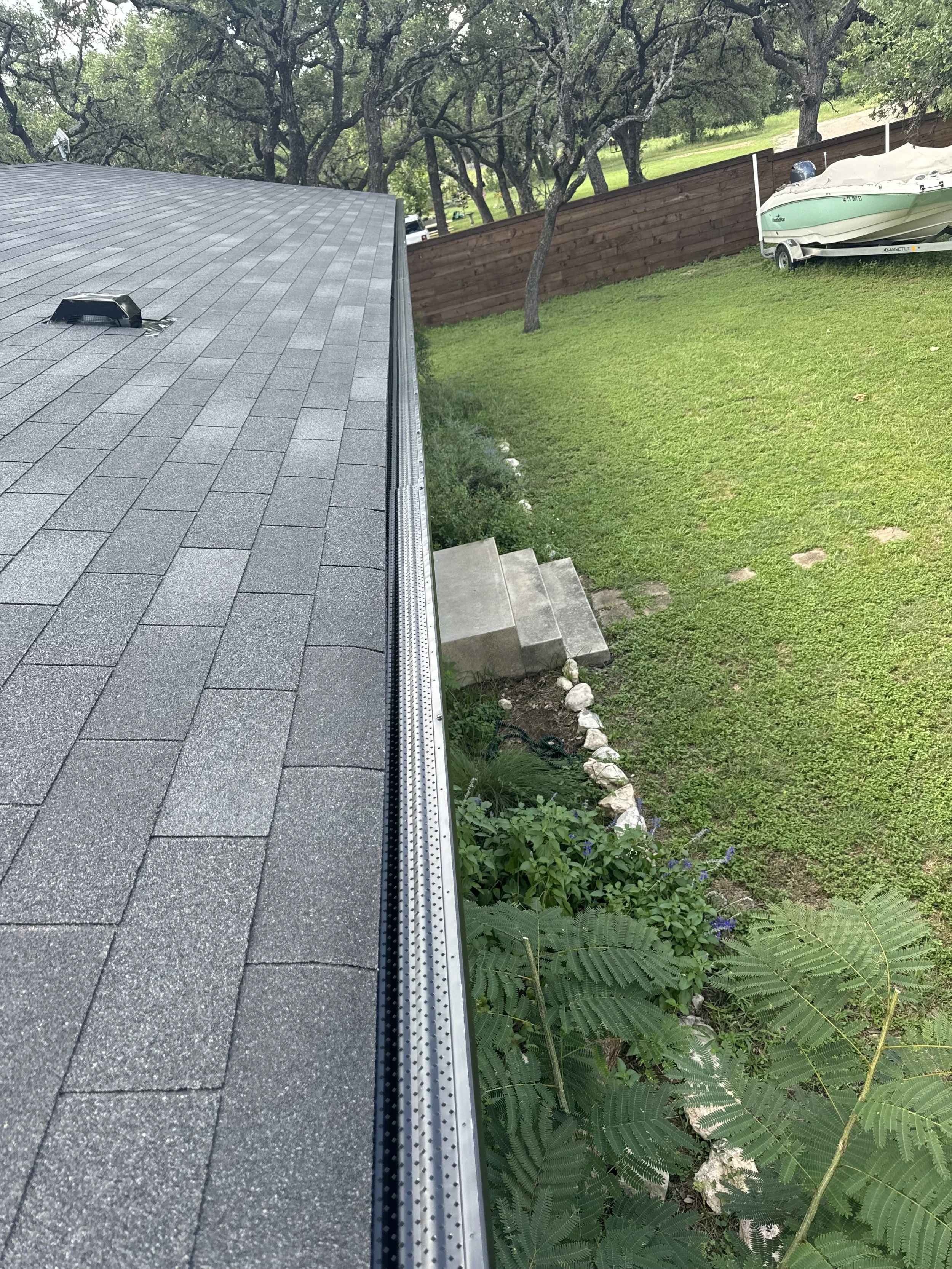 A rooftop with gray asphalt shingles and a metal gutter system, overlooking a garden with green plants and a set of concrete steps leading down to the lawn. In the background, there's a boat on a trailer, trees, and a wooden fence.