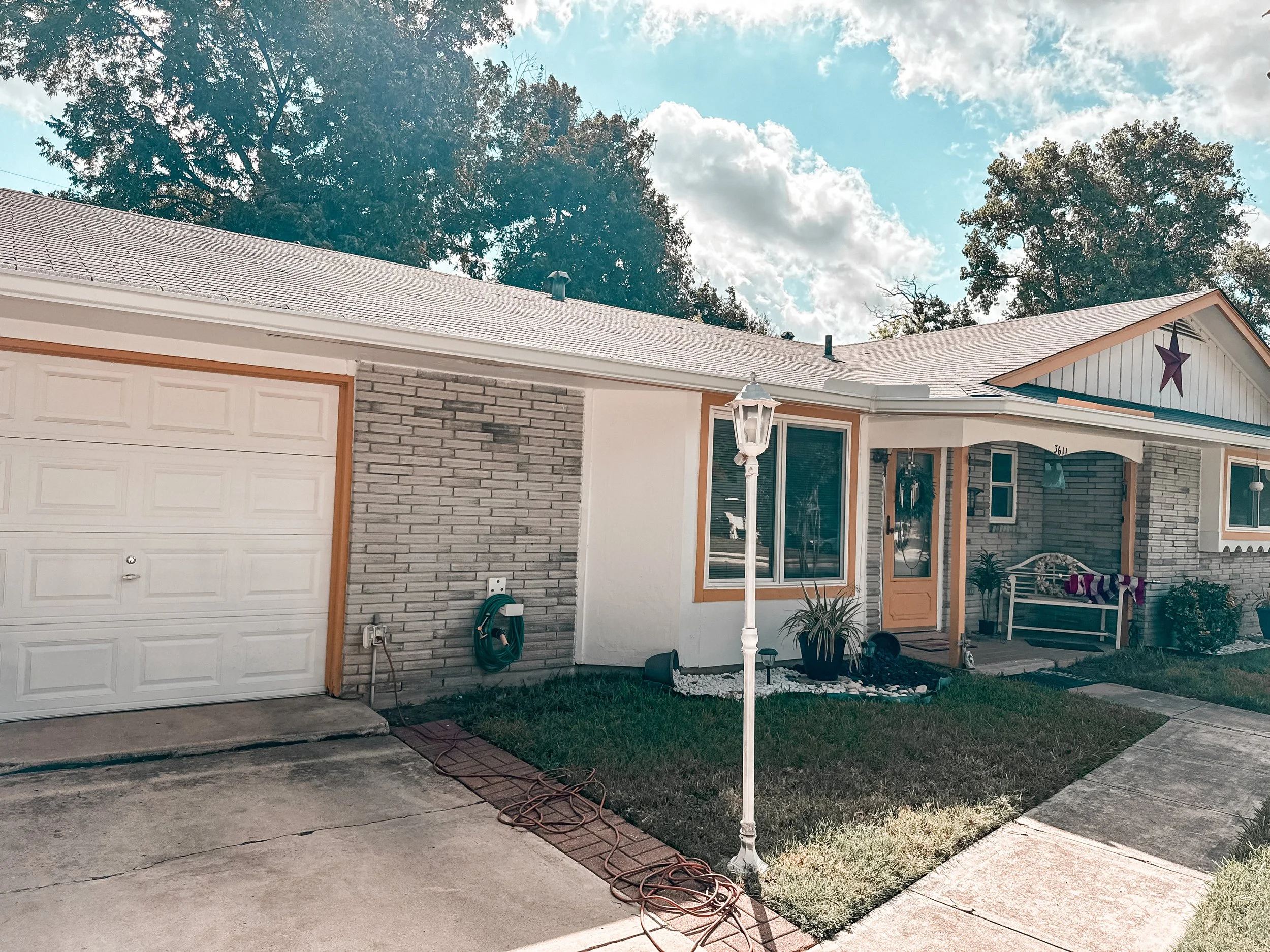 A single-story house with a brick and white exterior, a closed garage door, a small front yard with a lamp post, plants, and a sidewalk. Cloudy sky with some blue visible.