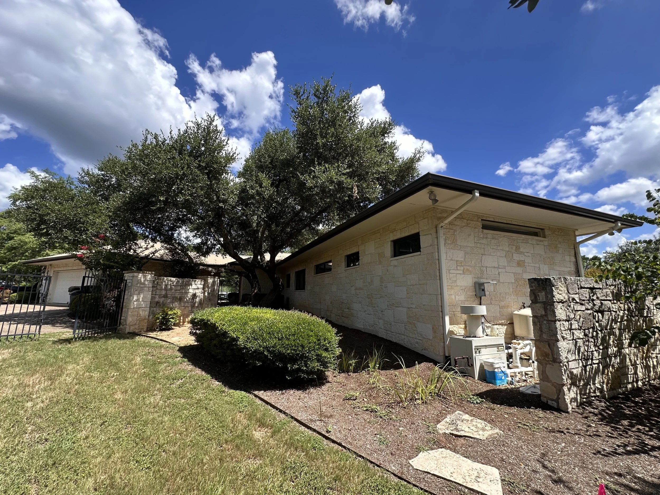 View of a house with a stone exterior wall, large tree, and a blue sky with clouds.