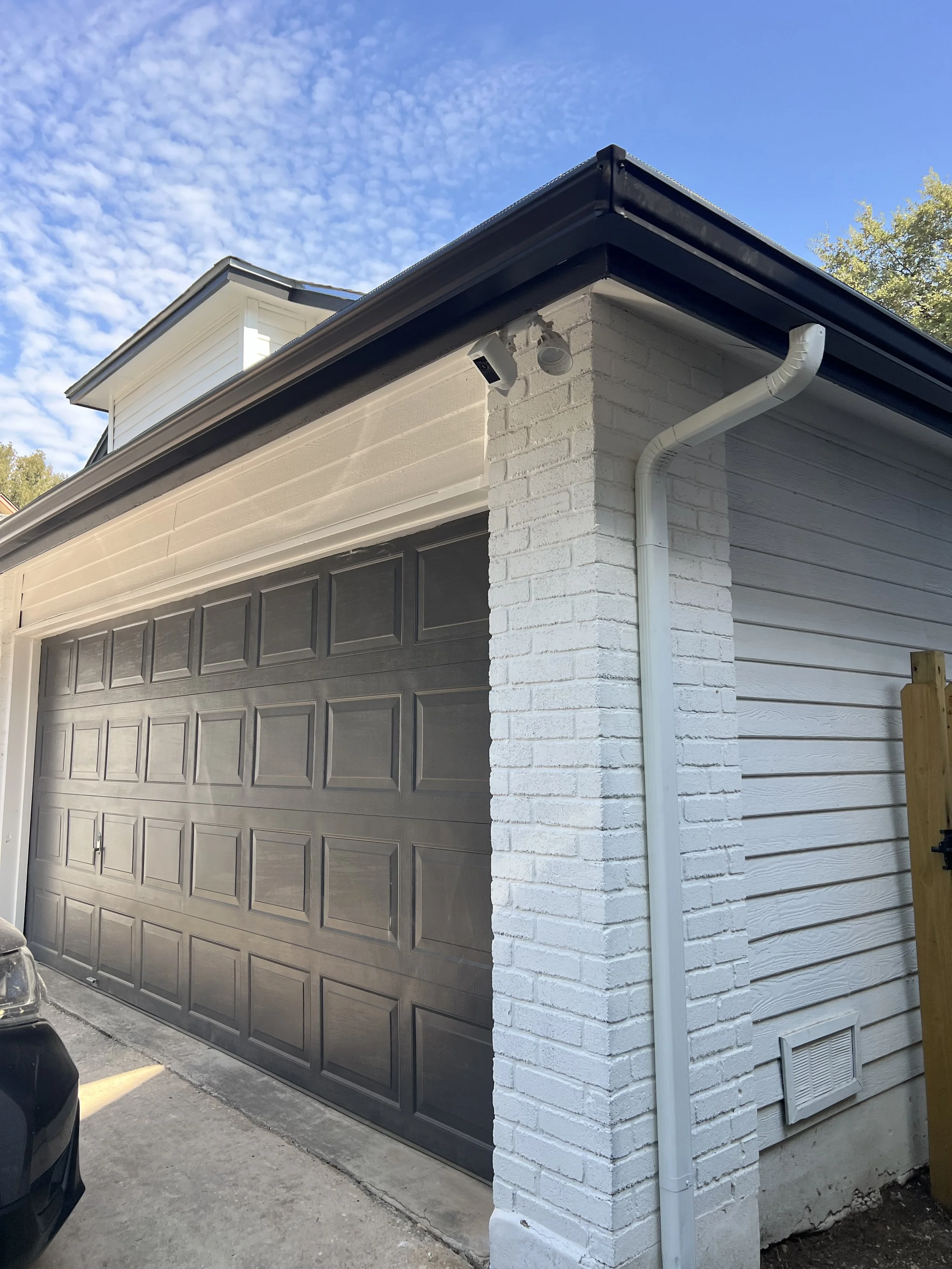 Exterior view of a house garage with a black sectional garage door, white brick wall, white horizontal siding, gutter system, and security camera, with blue sky and trees in the background.