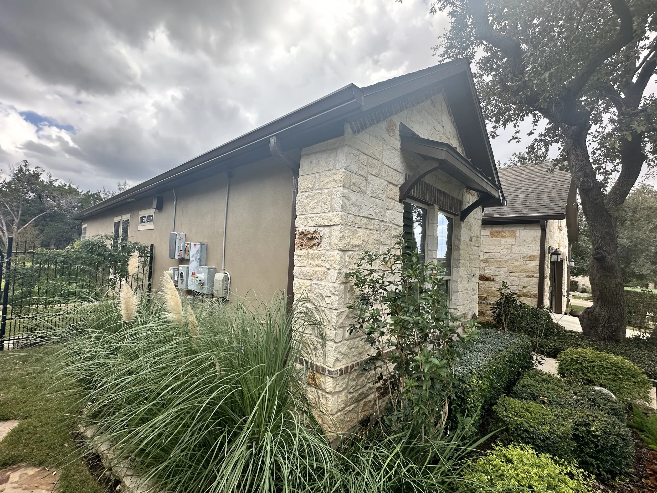Side view of a house with a stone and stucco exterior, surrounded by landscaping, trees, and cloudy sky.