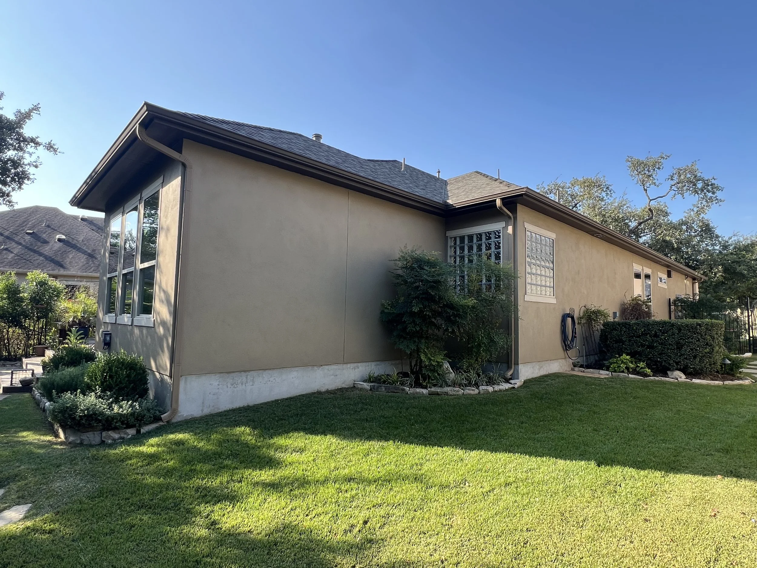 Side view of a beige house with a well-maintained green lawn, bushes, and trees under a clear blue sky.