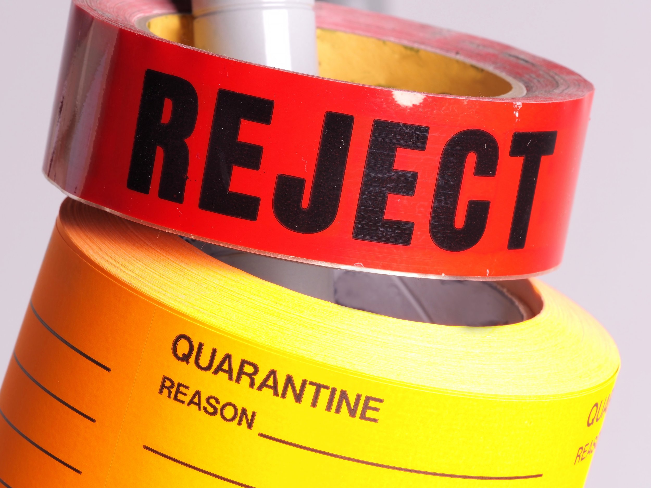 Close-up of a red adhesive tape with the word 'REJECT' in black letters above a yellow quarantine label with blank fields for 'REASON' and 'QUARANTINE' on a gray background.
