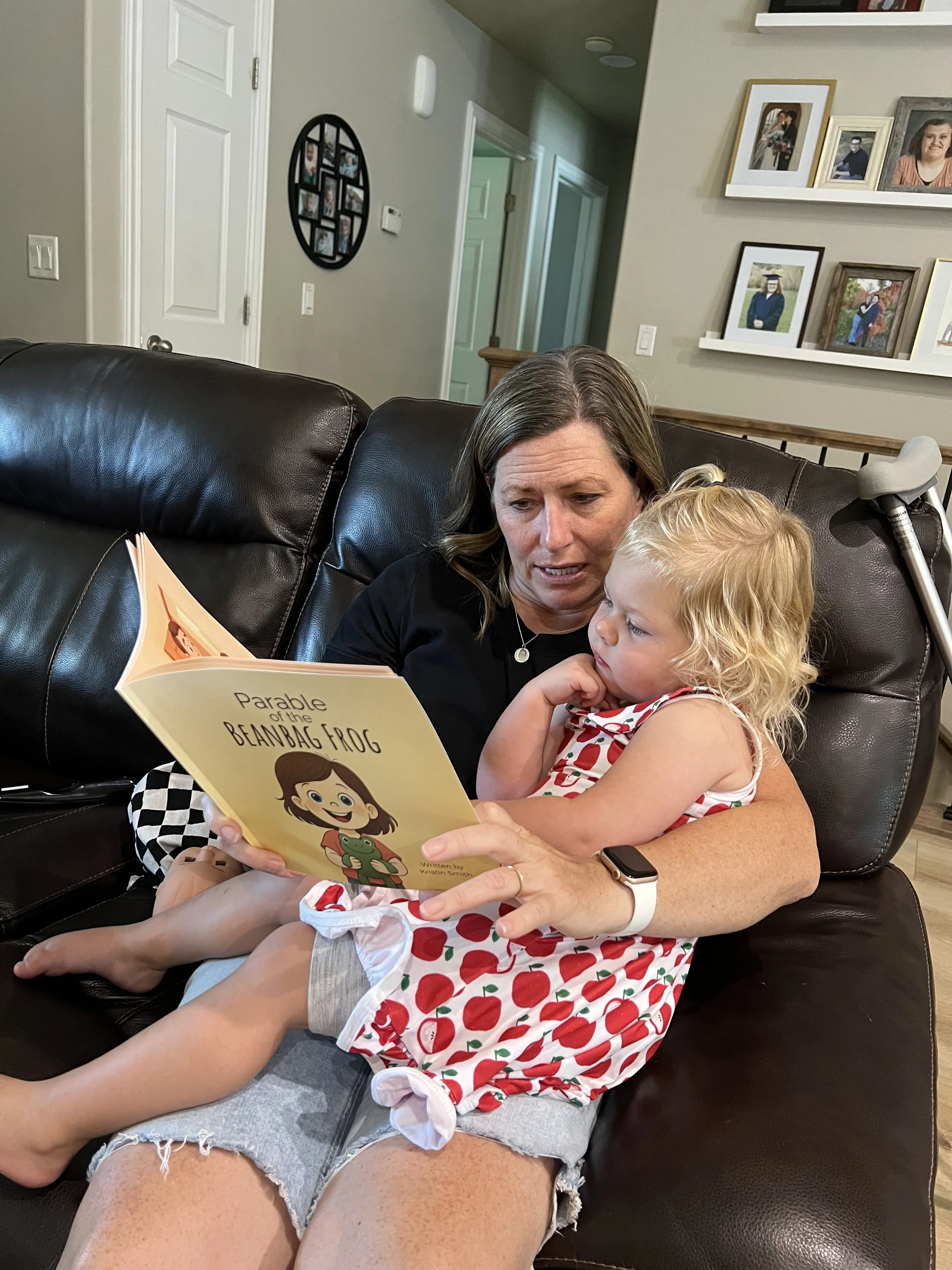 A woman and a young girl sitting on a black leather couch, reading a children's book titled 'Parable of the Beanbag Frog.' The woman appears to be engaged and the girl is listening attentively. The woman is wearing a smartwatch and a black shirt, and the girl is in a red and white polka dot dress. In the background, there are photo frames on a wall and an open doorway.