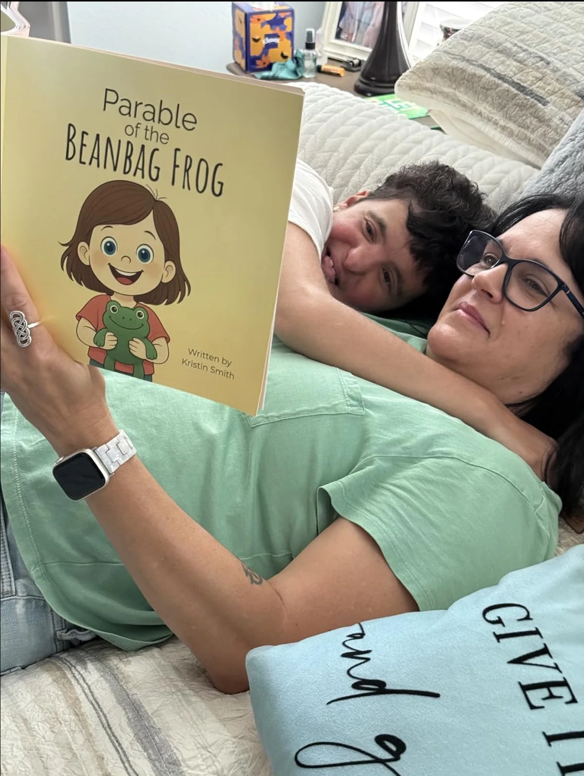 A woman with glasses lying on a couch, reading a children's book titled "Parable of the Beanbag Frog" to a young boy who is lying next to her. The woman is wearing a light green shirt and a smartwatch, and the boy is smiling and has dark, curly hair.