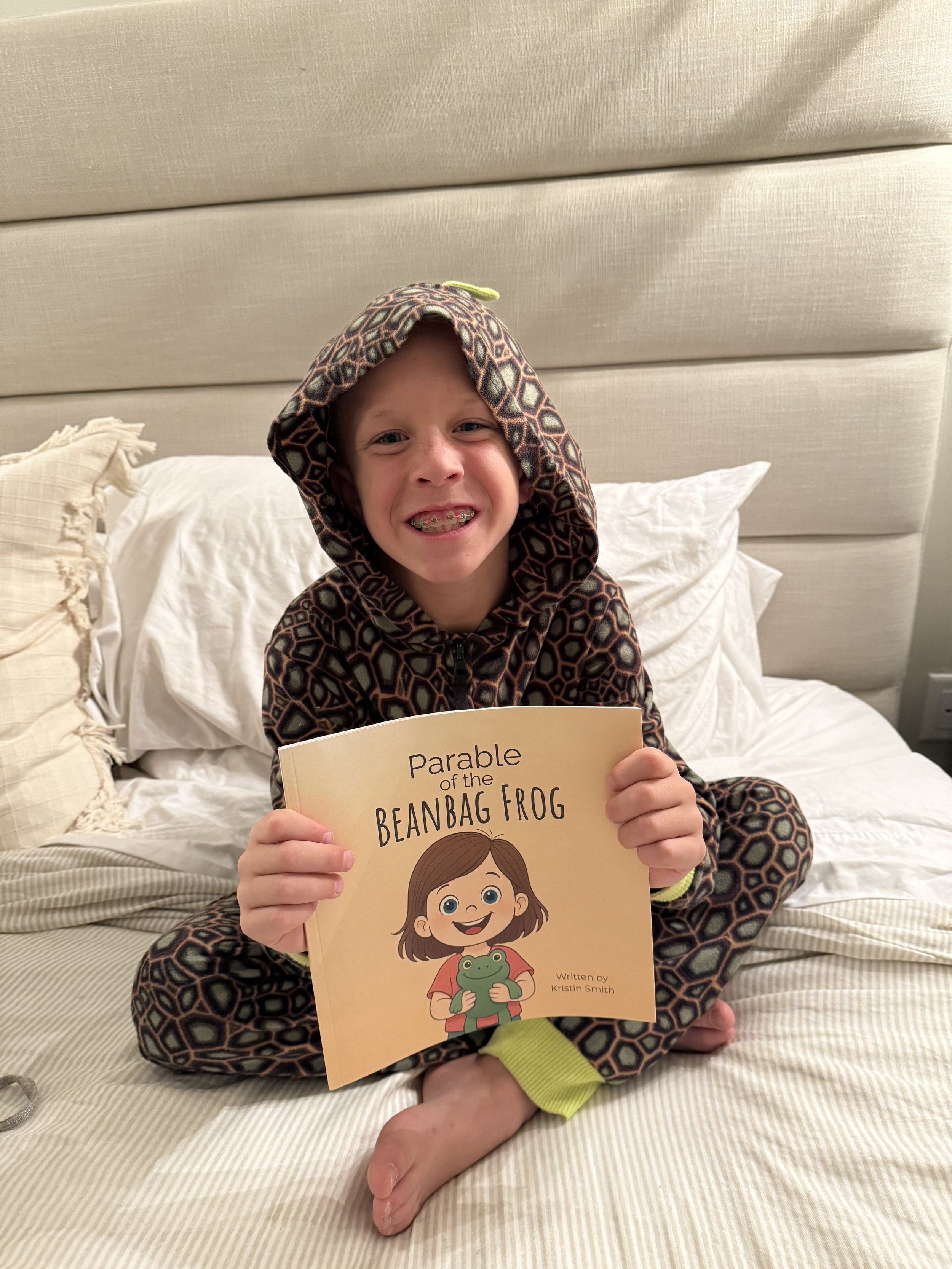 Young girl sitting on a bed, holding a children's book titled 'Parable of the Beanbag Frog', wearing a hoodie with a geometric pattern, smiling.