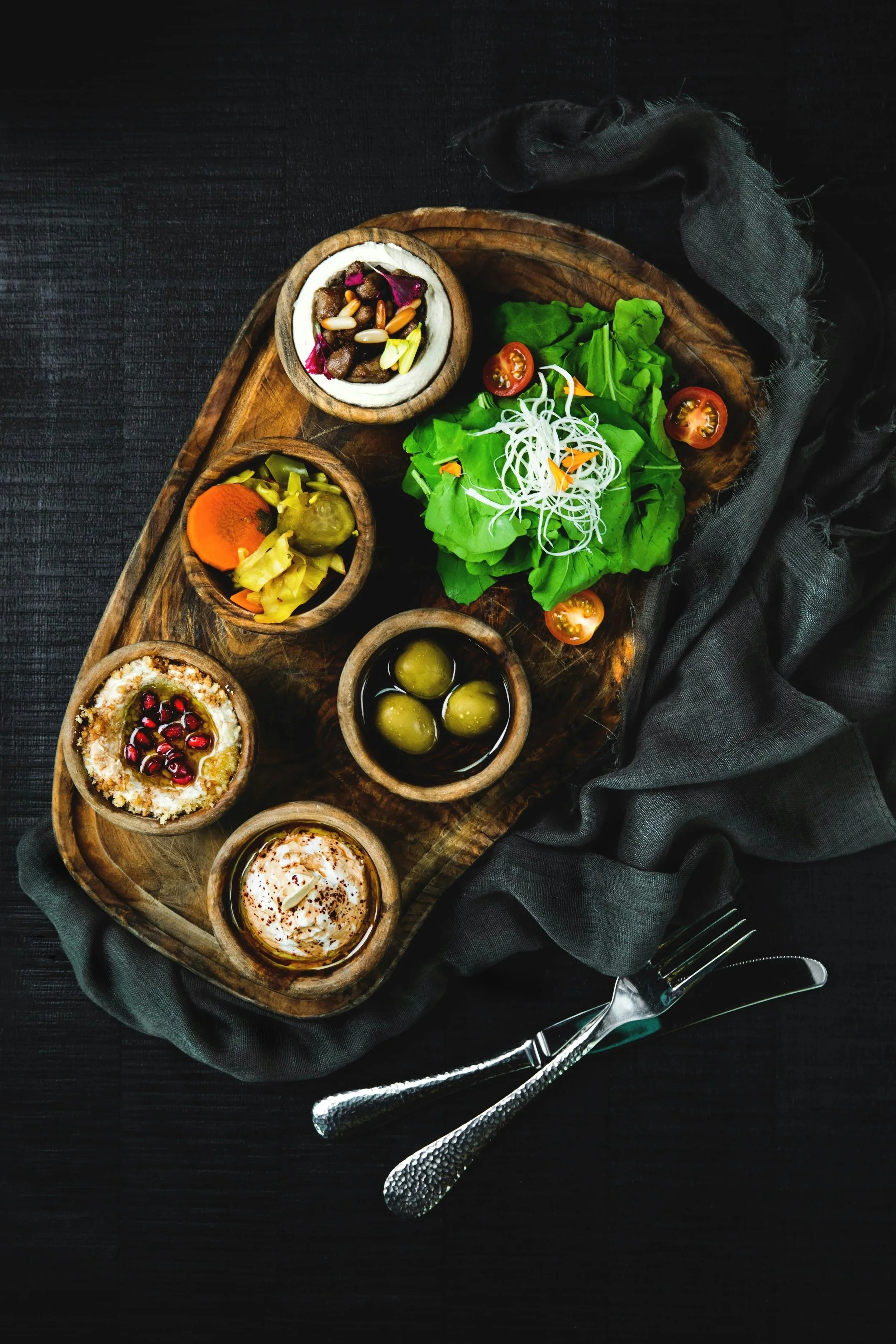 Verschiedene mediterrane Vorspeisen und Salate auf einem Holztablett, serviert mit Besteck und dunklem Tuch