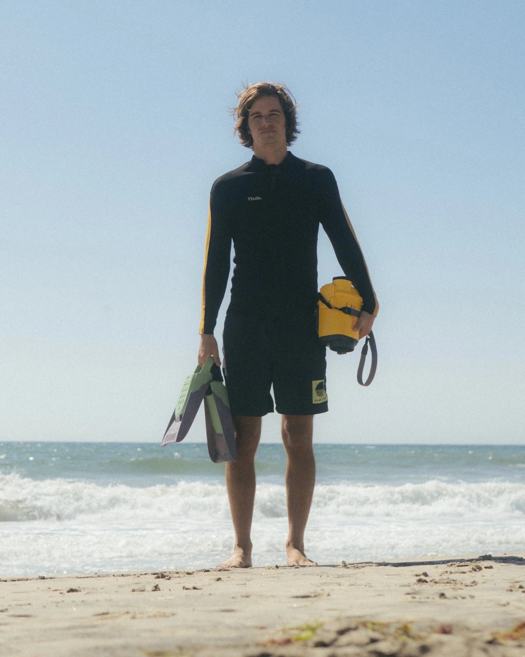 Young man standing barefoot on the beach holding swim fins and a yellow underwater camera, dressed in a black wetsuit with yellow accents.
