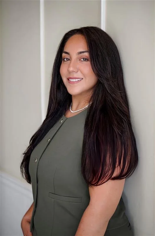 A woman with long dark hair, wearing a green top, pearl necklace, standing against a white wall, smiling at the camera.