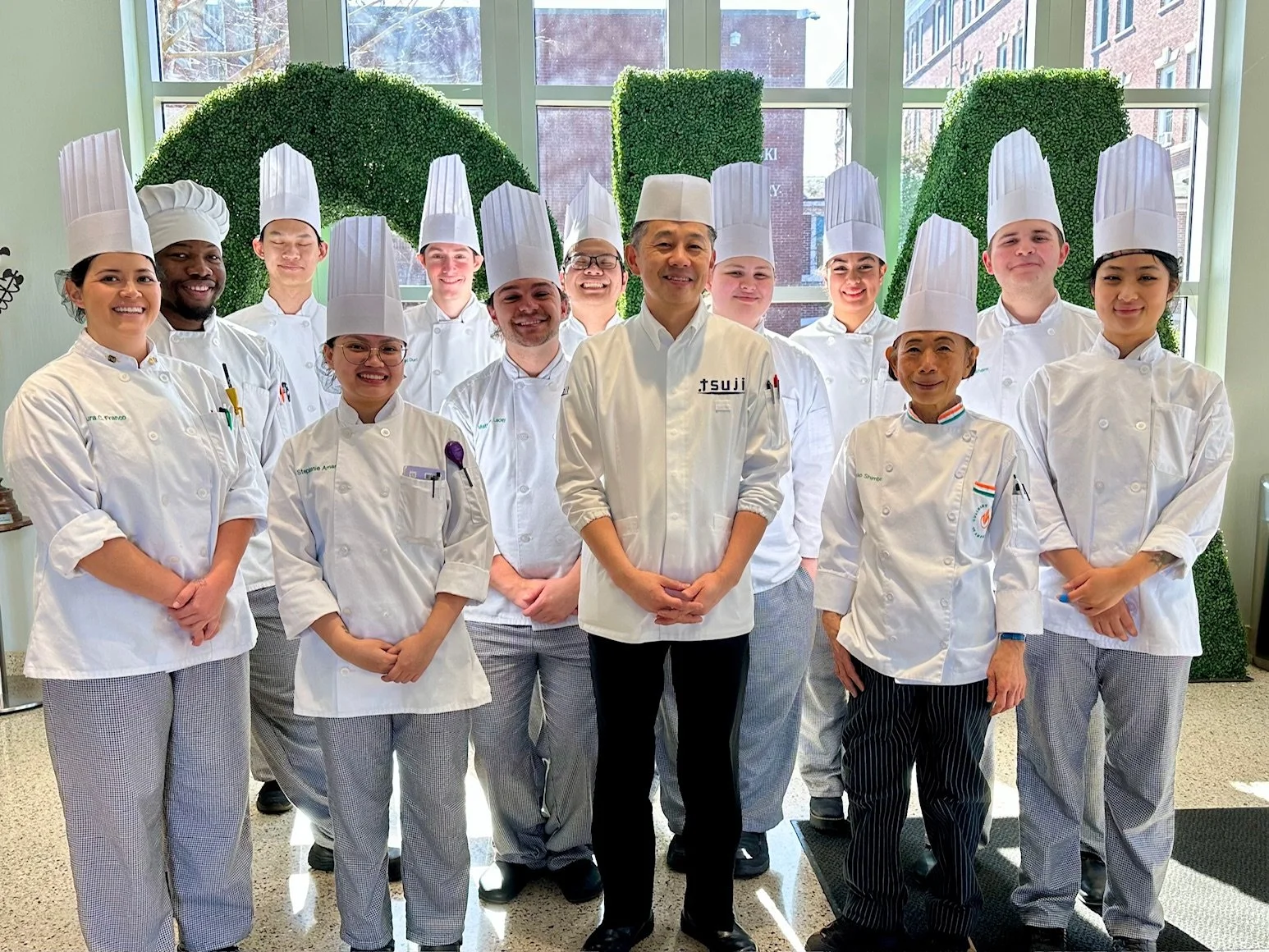 Group of chefs in white uniforms and tall hats standing in a bright kitchen and smiling.