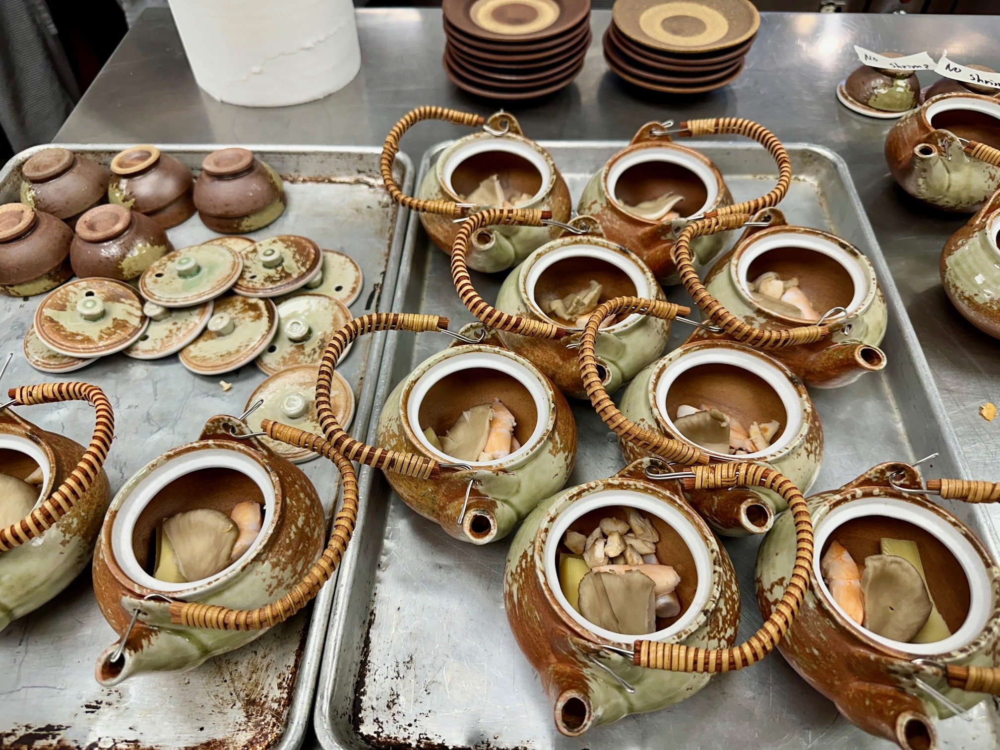 Clay teapots and cups filled with tea, with some sliced ginger and other ingredients inside, arranged on metal trays on a stainless steel counter. There are also stacks of small bowls and lids nearby.