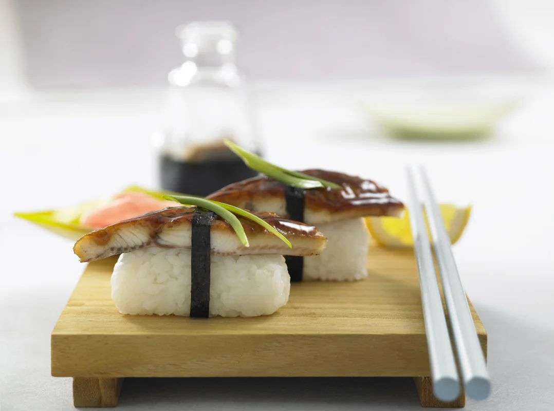 Two pieces of unagi nigiri sushi on a wooden board, garnished with green onion and a black strip of seaweed, with a bottle of soy sauce in the background.