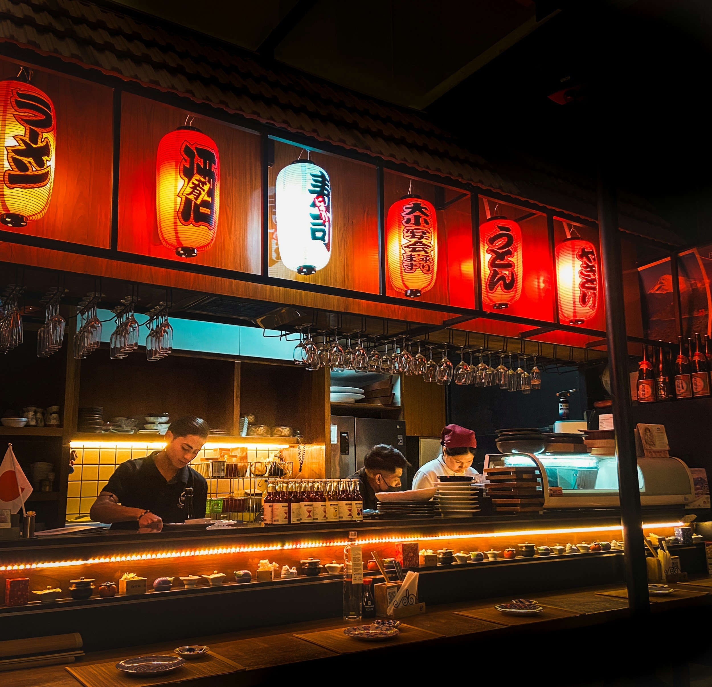 Interior of a Japanese restaurant with hanging red and white lanterns and two chefs working behind a sushi bar.
