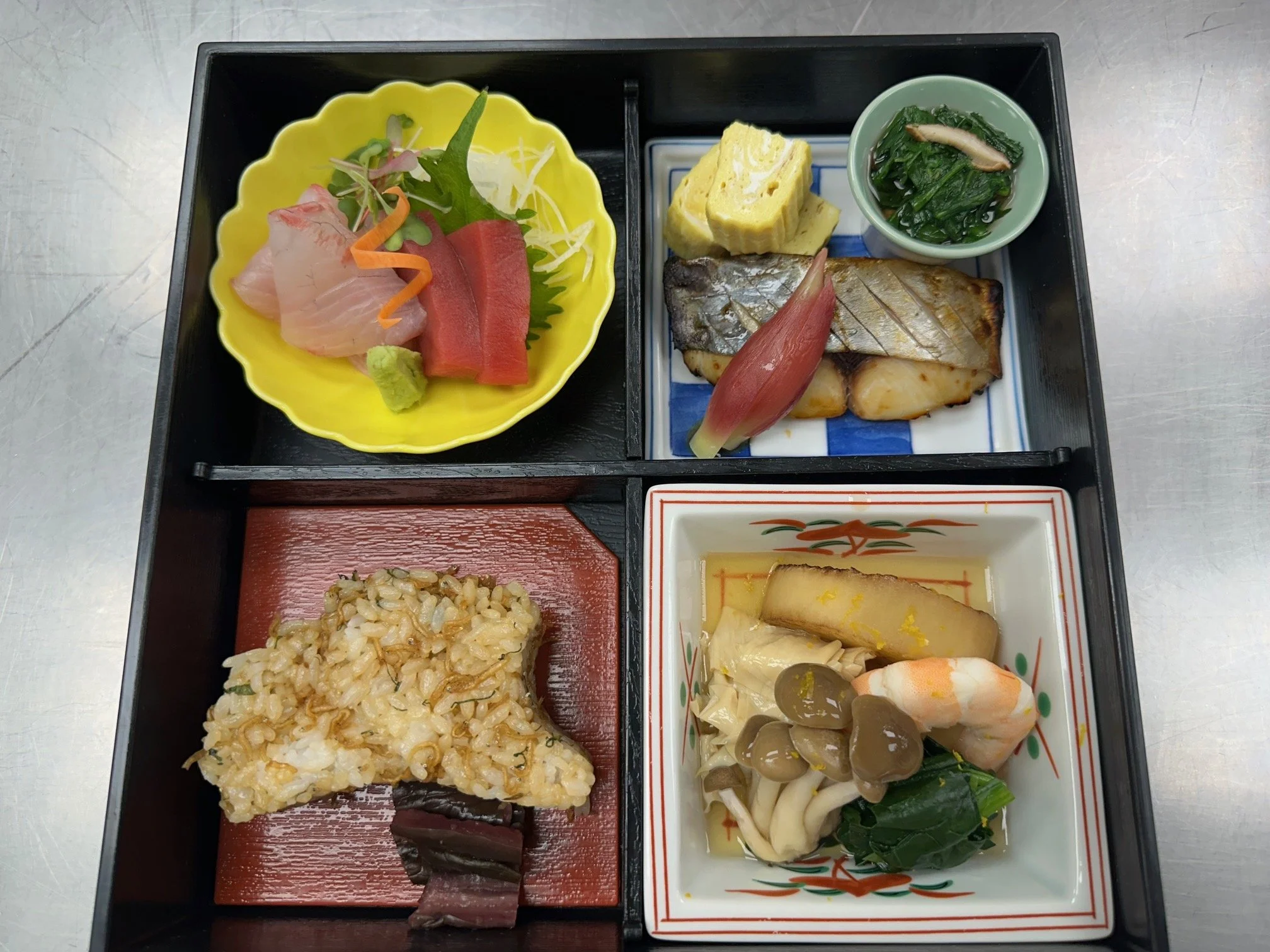 A Japanese bento box with sashimi, grilled fish, tamagoyaki, vegetables, rice, and pickles.