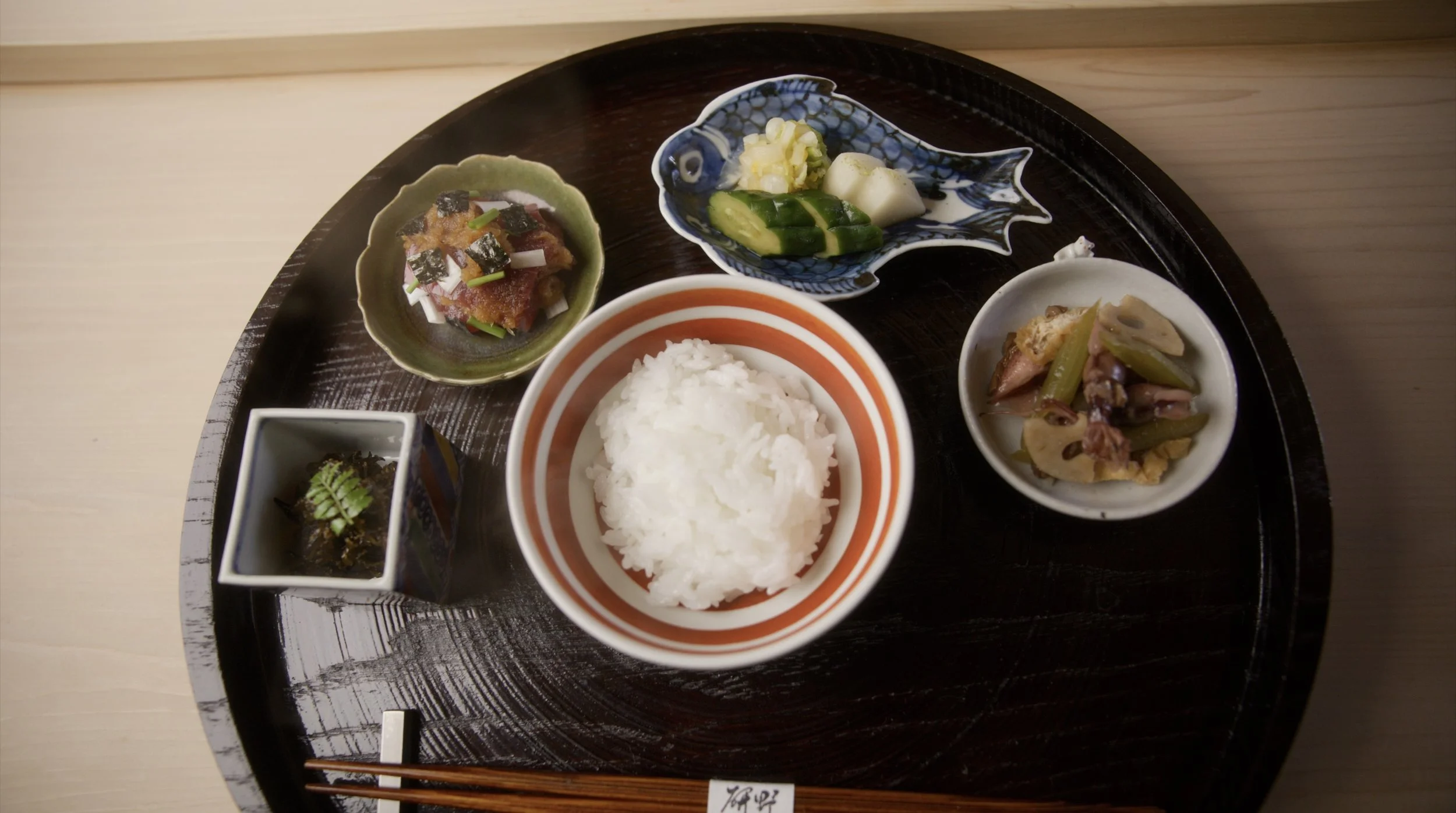 Traditional Japanese meal served on a black round tray includes a bowl of white rice, small dishes with pickled vegetables, seaweed, and simmered vegetables, along with leafy greens, and garnishes.