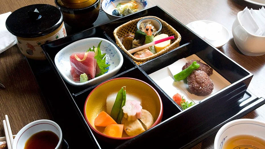 Traditional Japanese bento box with sashimi, pickled vegetables, grilled meat, and side dishes, served with bowls of soup and tea.