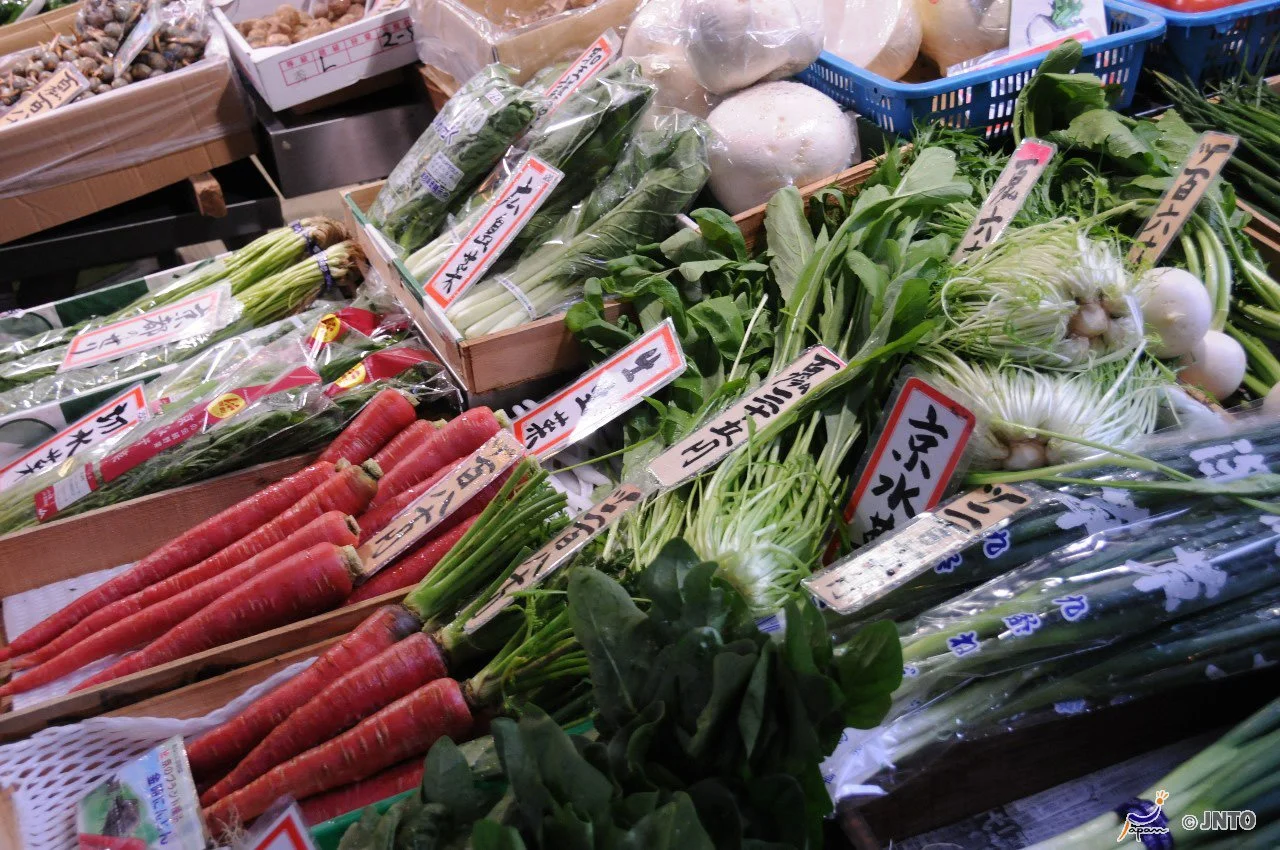 Fresh vegetables including carrots, green onions, and leafy greens on display at a market stall with handwritten signs in Japanese.