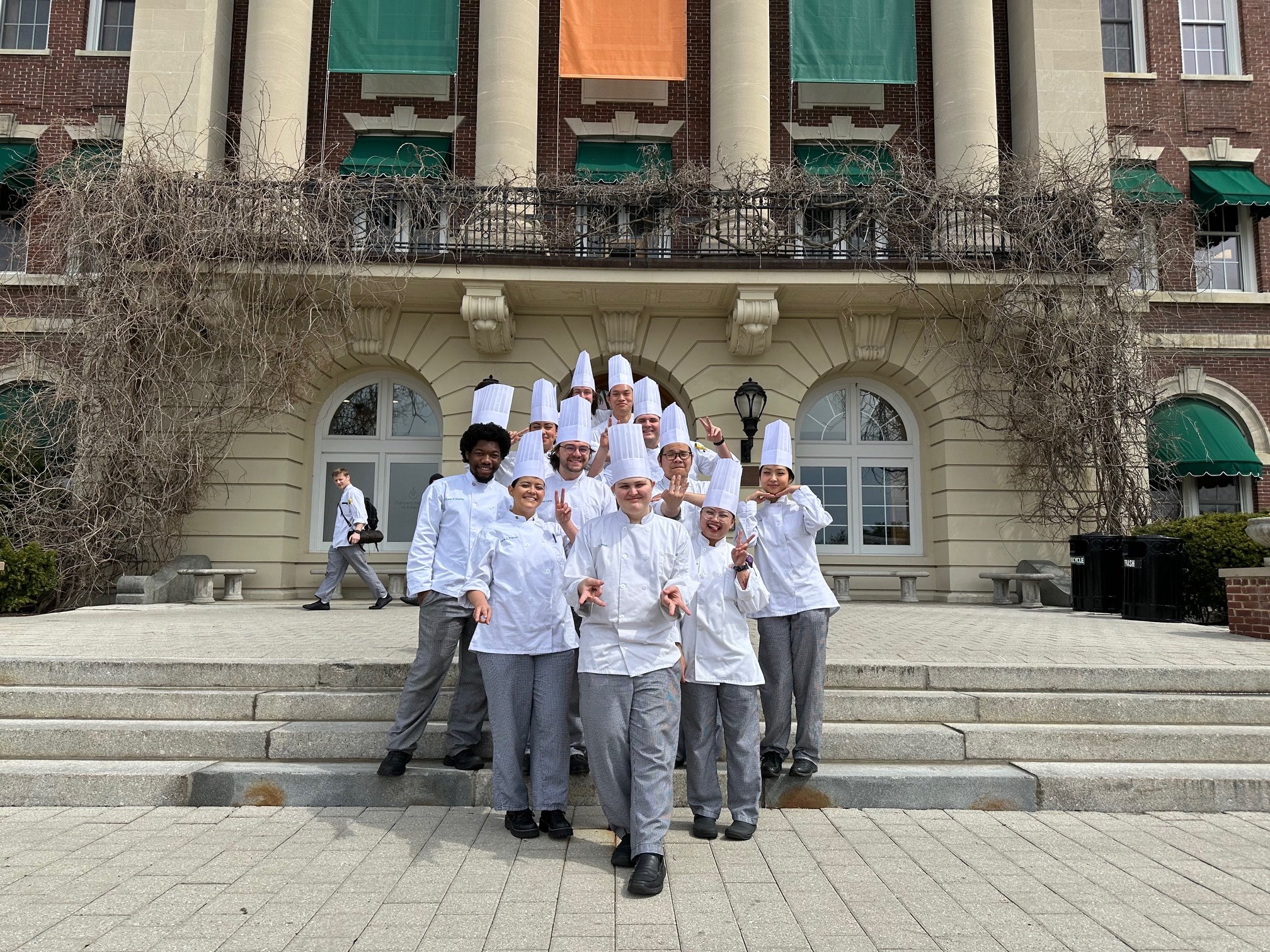 Group of chefs in white uniforms and tall hats posing on steps outside an elegant building with arched windows, some making peace signs and smiling.
