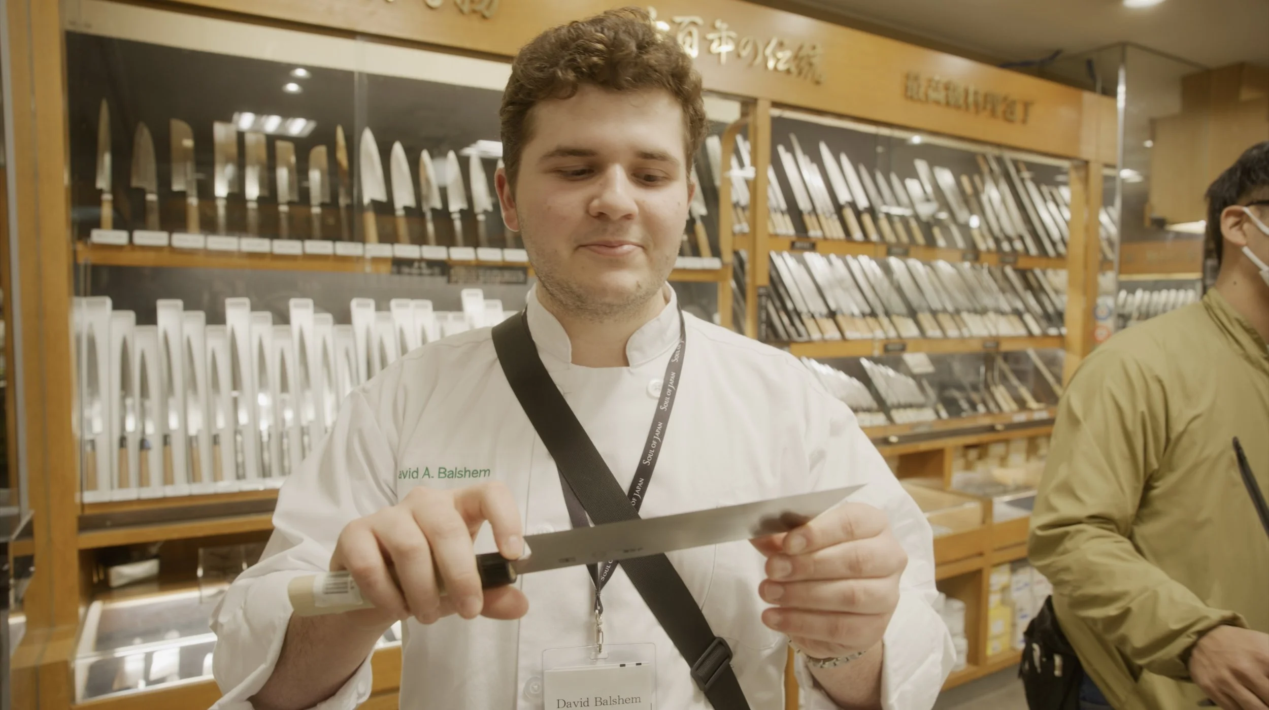 A young man in chef attire with a name tag reading David Balshem is holding and examining a kitchen knife in a store that sells knives, with shelves of knives displayed behind him.