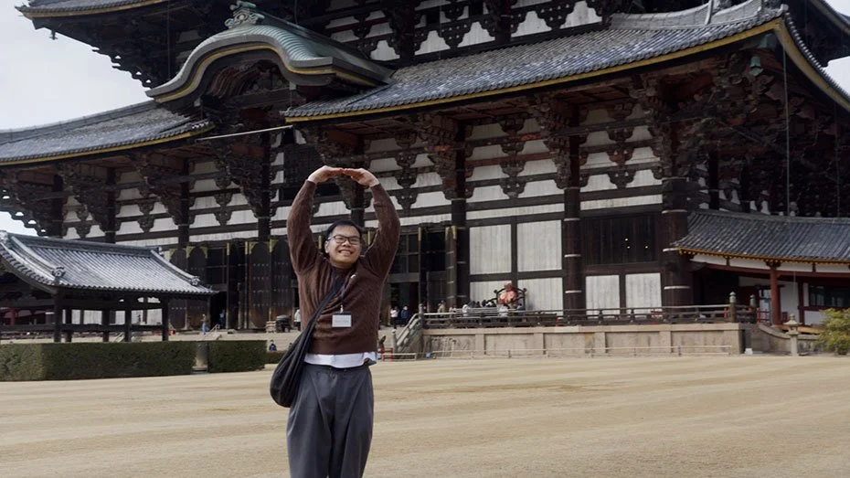 Person standing in front of a traditional Japanese temple, smiling, raising arms above head in a circular shape.