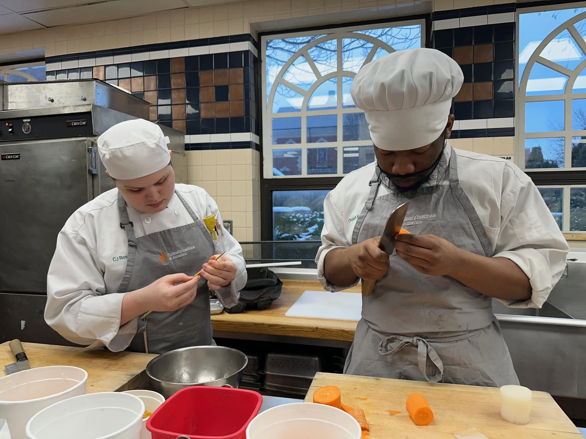 Two culinary students in chef hats and aprons preparing vegetables in a professional kitchen.