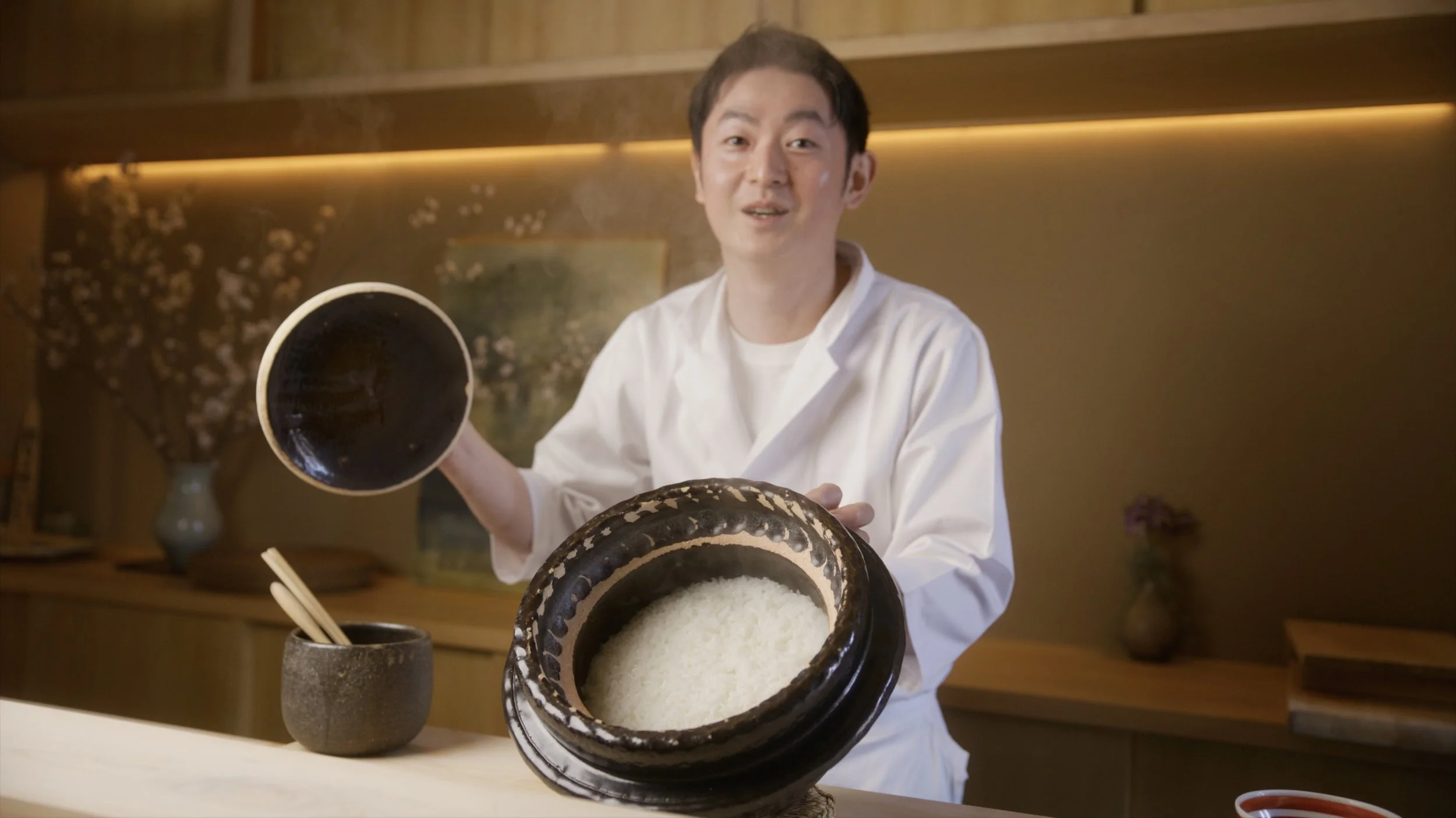 Chef in white coat holding a bowl of cooked rice with a smiling expression, in a kitchen setting.