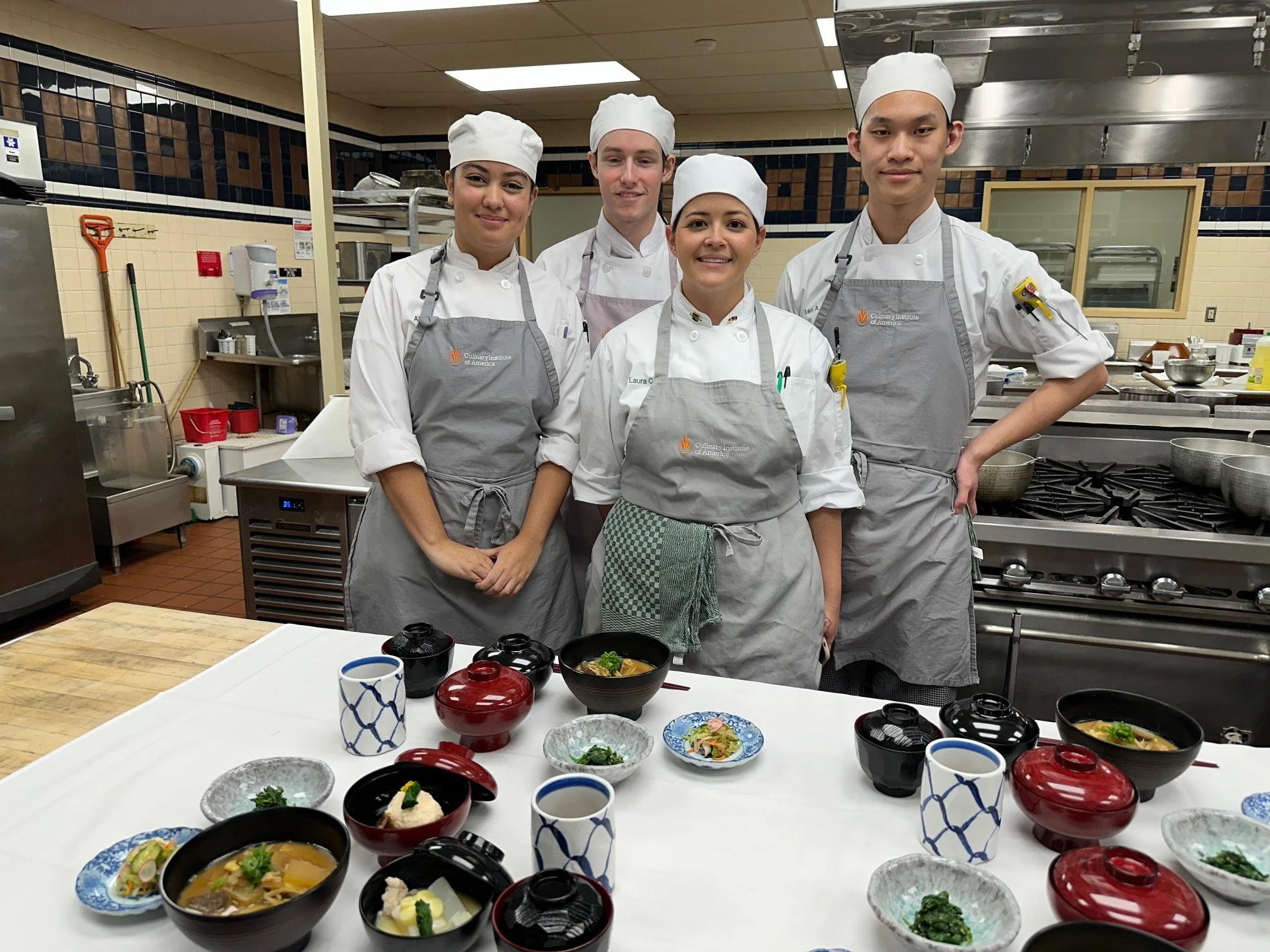 Four culinary students in chef uniforms and aprons standing behind a table with Japanese dishes, in a professional kitchen.
