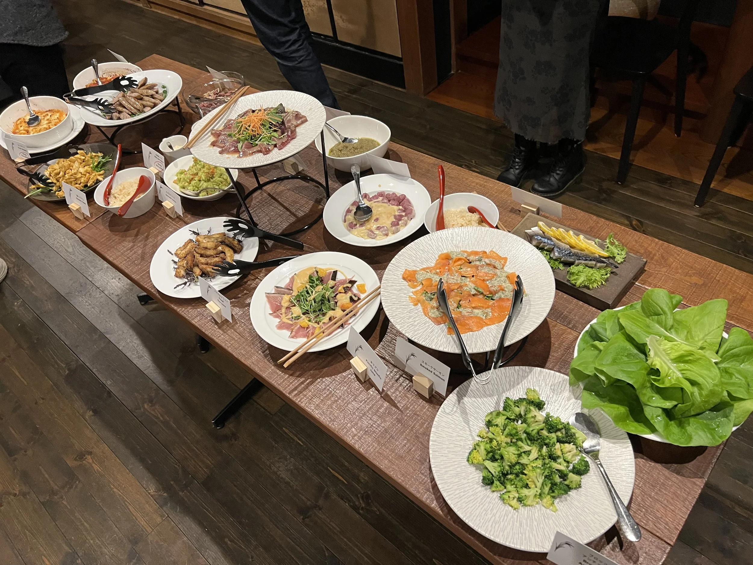 A table filled with various dishes including sashimi, salads, chicken wings, and other appetizers, with small labels in front of each dish, and a large bowl of leafy greens at the side.
