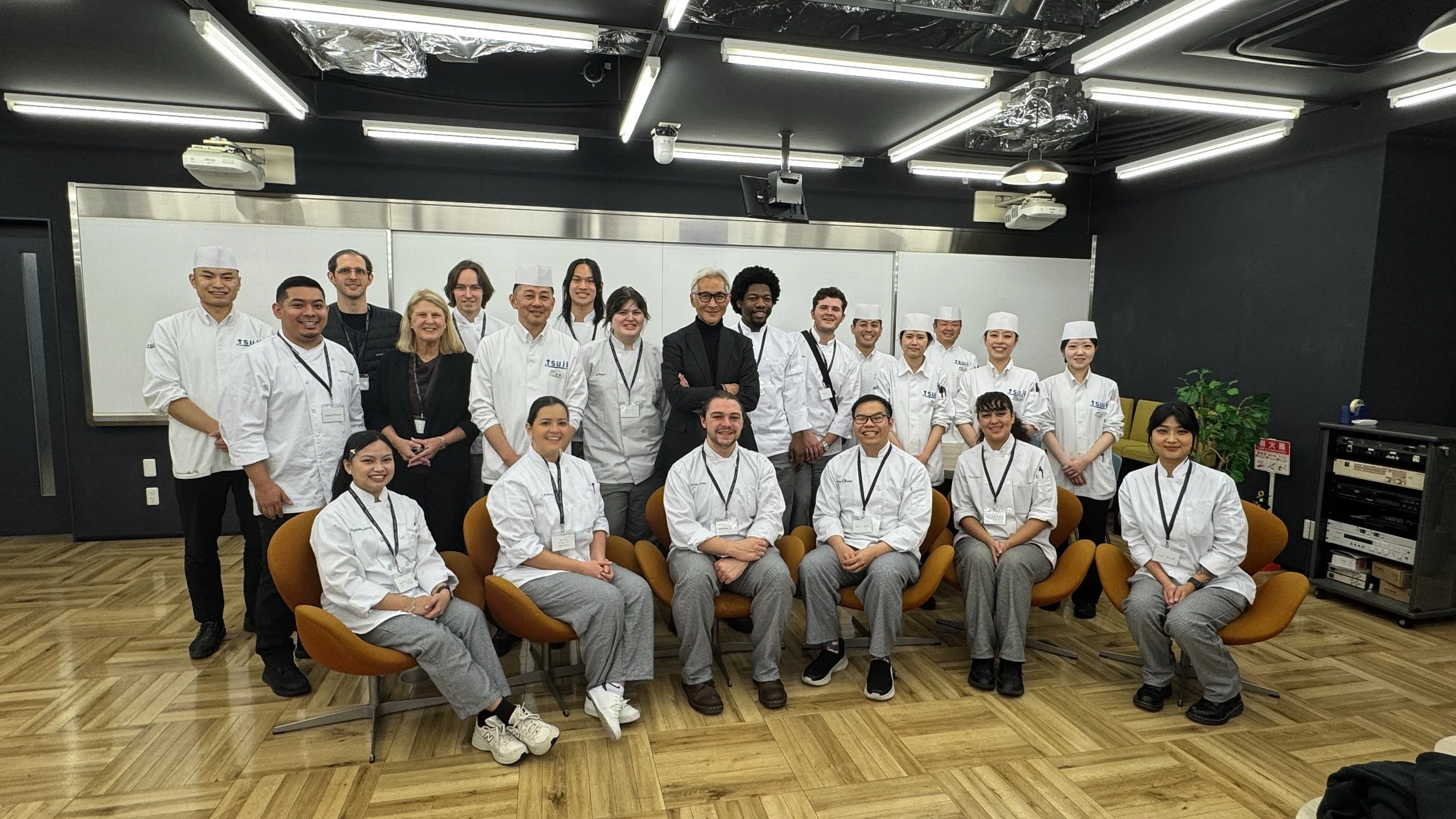 A group photo of twenty-one people, including students in chef uniforms and instructors, in a classroom or conference room setting with whiteboards and a wooden floor.