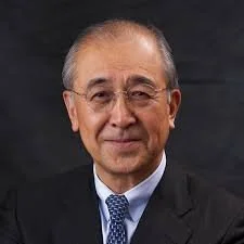 Portrait of an older man wearing glasses, a dark suit, a white shirt, and a blue tie, smiling at the camera against a dark background.