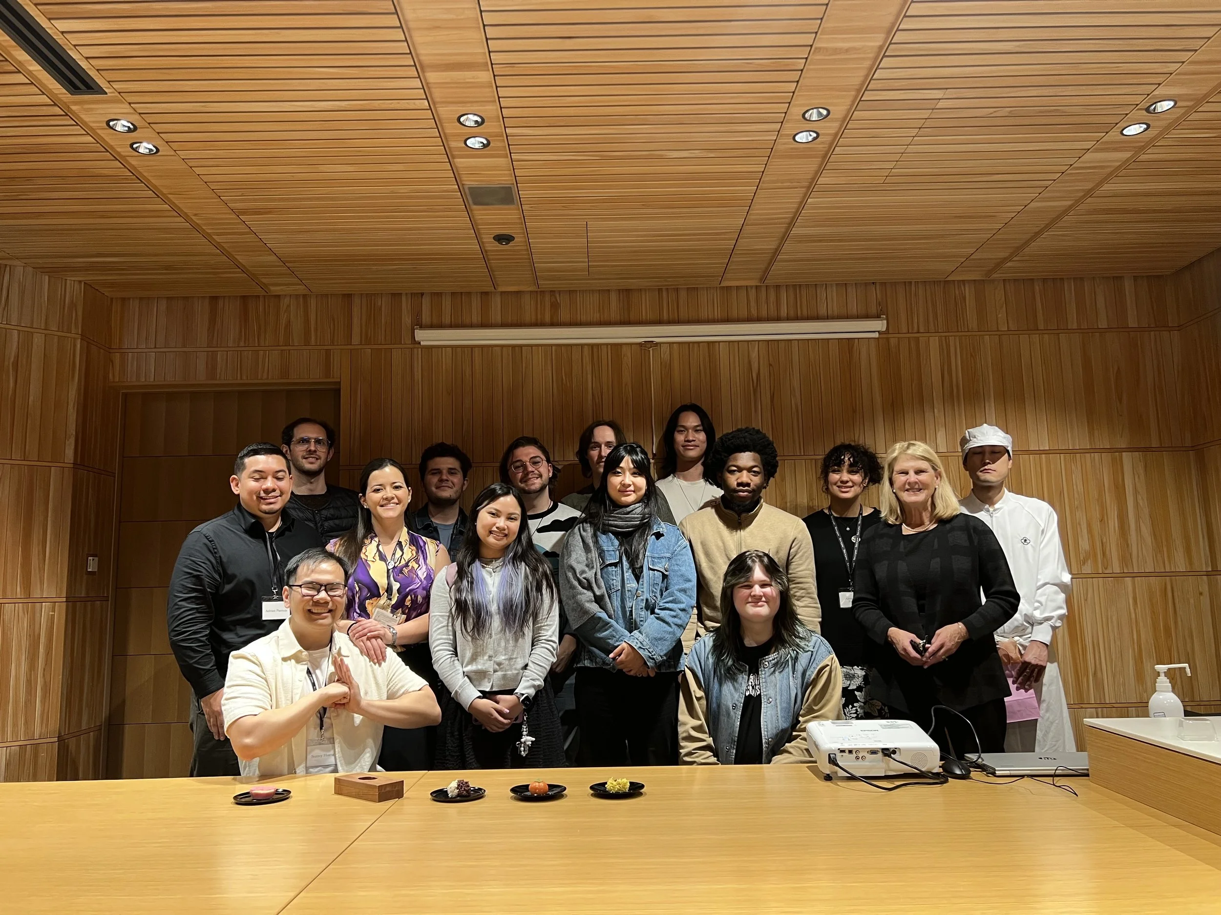 Group of 15 people posing in a wood-paneled conference room, some wearing conference badges, with a projector and snacks on the table in front.