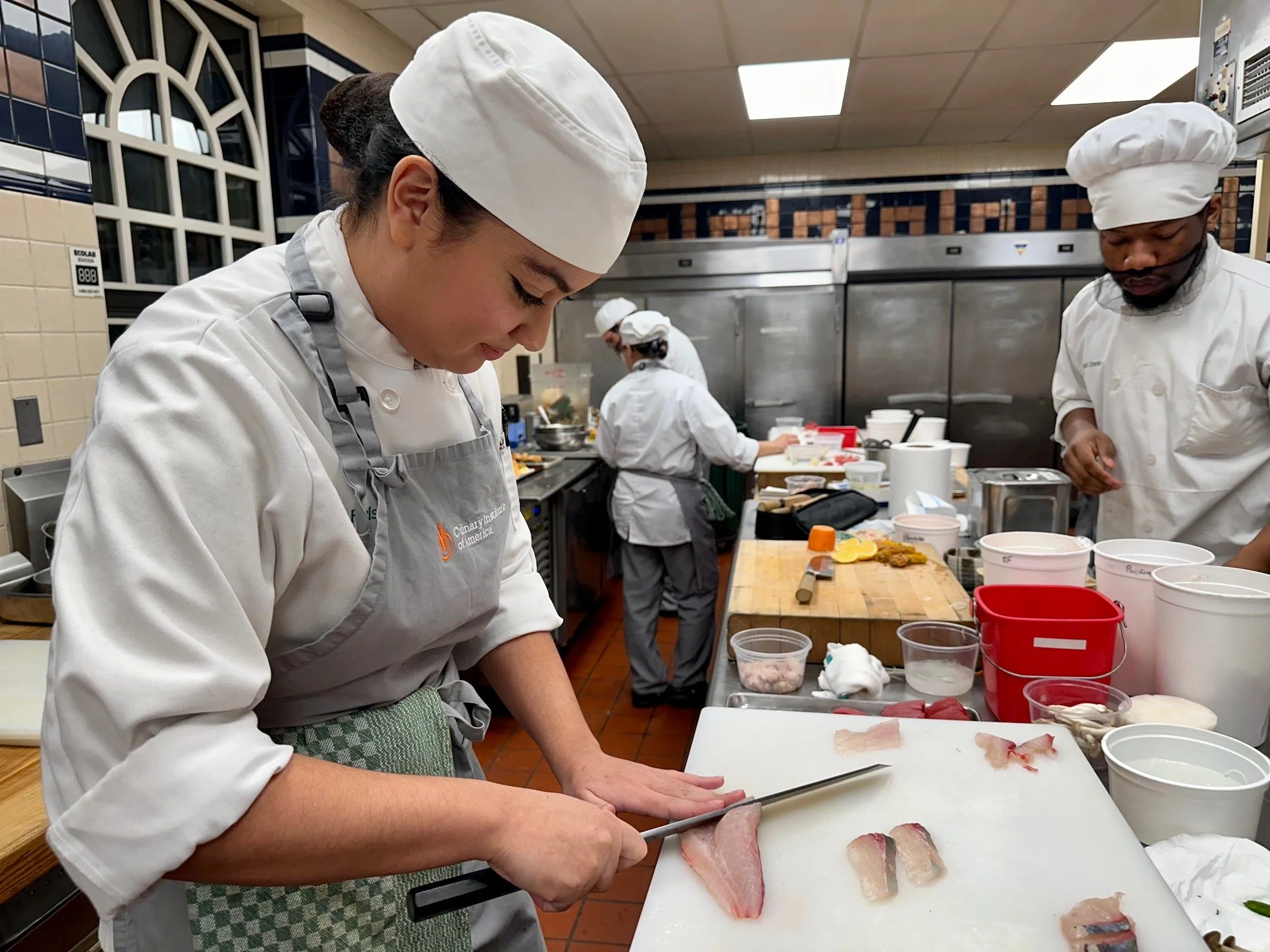 Chef chopping fish fillets in a professional kitchen with other chefs preparing food in the background.