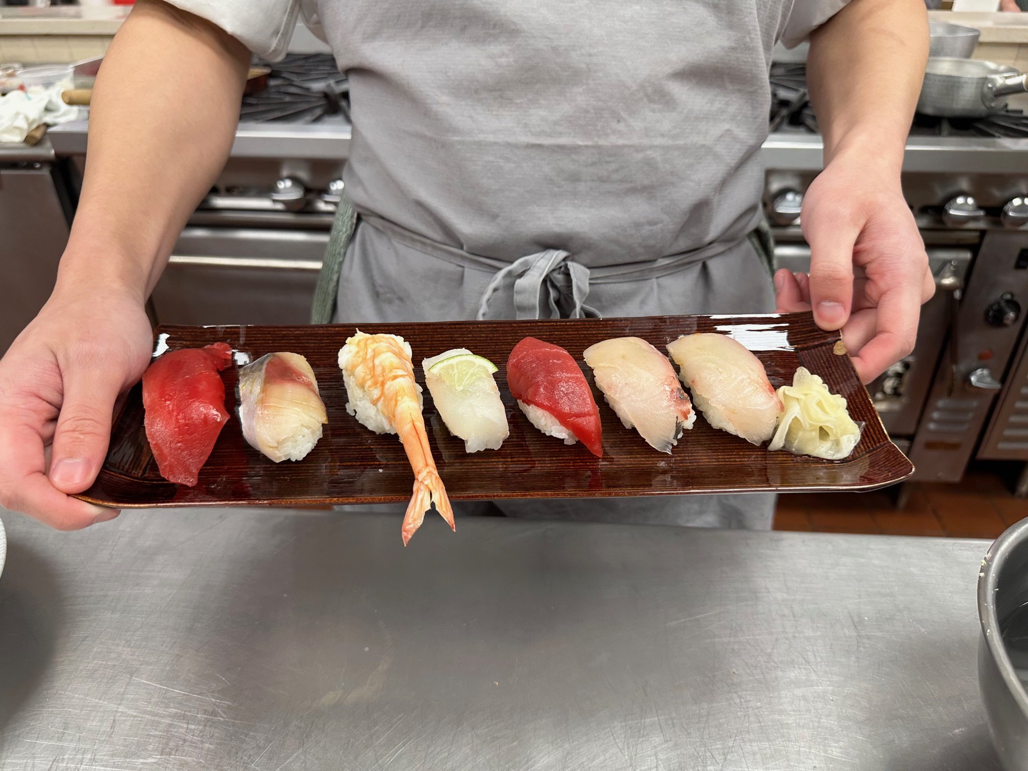 Person holding a rectangular wooden plate with assorted sushi, including slices of tuna, white fish, shrimp, and pickled ginger, in a kitchen setting.
