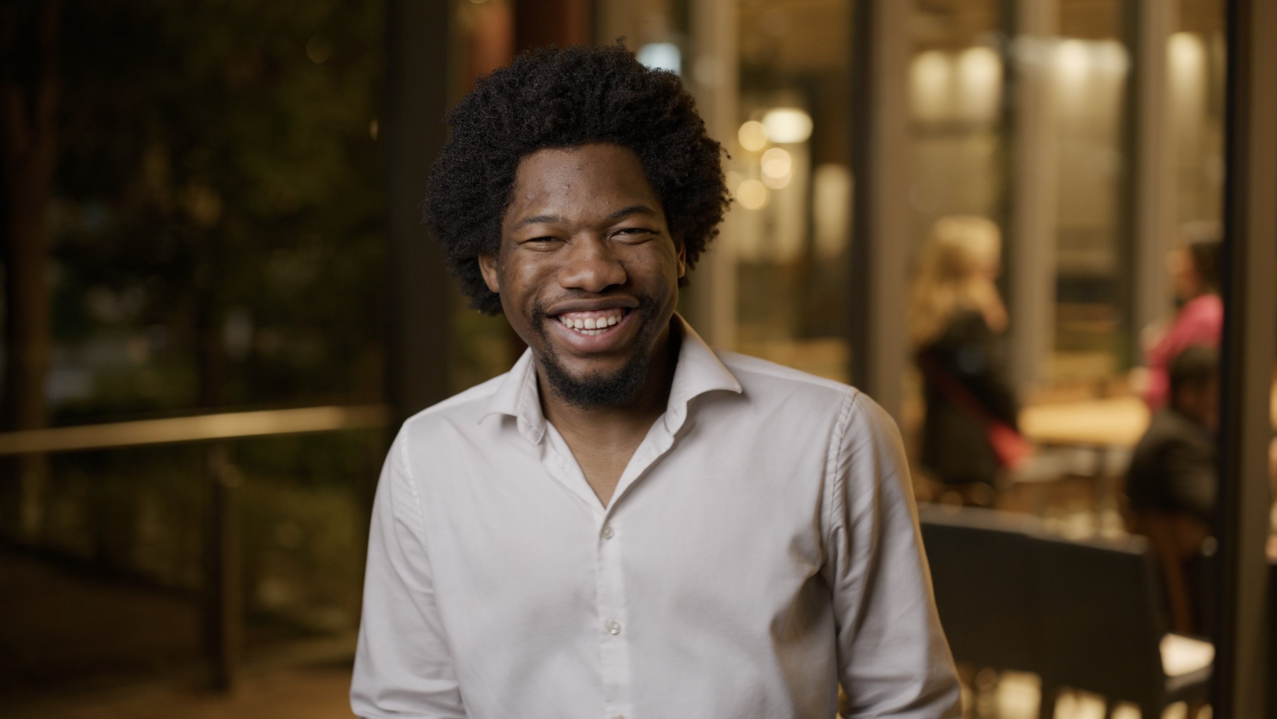 A smiling man with curly hair and a white shirt standing outdoors at night, with people sitting inside a building in the background.