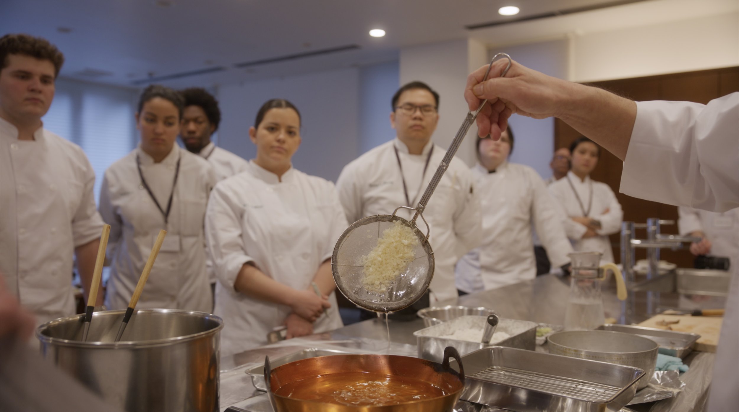 Culinary students in chef uniforms observing a chef straining cheese into a pot in a cooking class kitchen.
